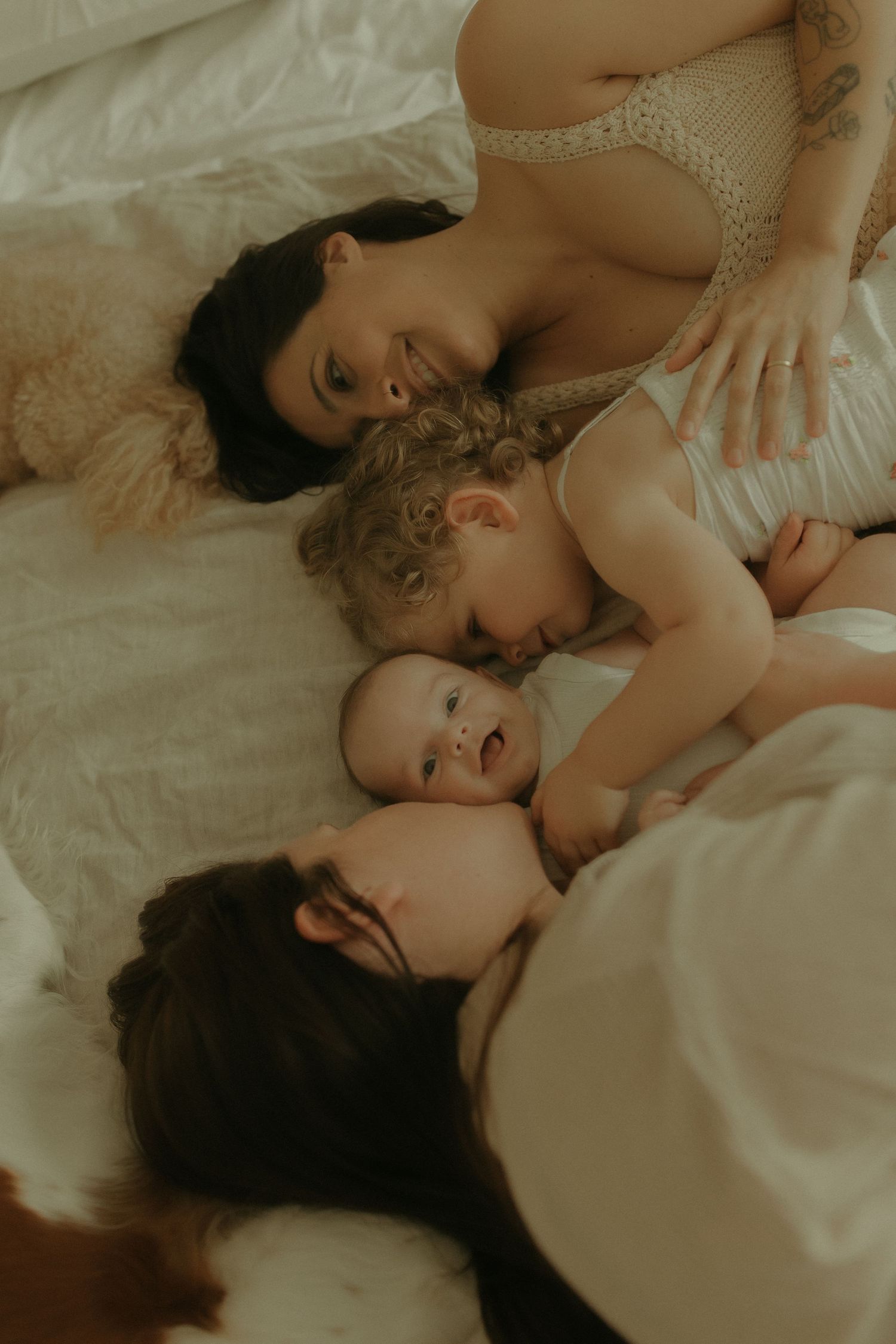 An intimate family portrait of adults and babies cuddling together on a cozy bed in warm lighting.