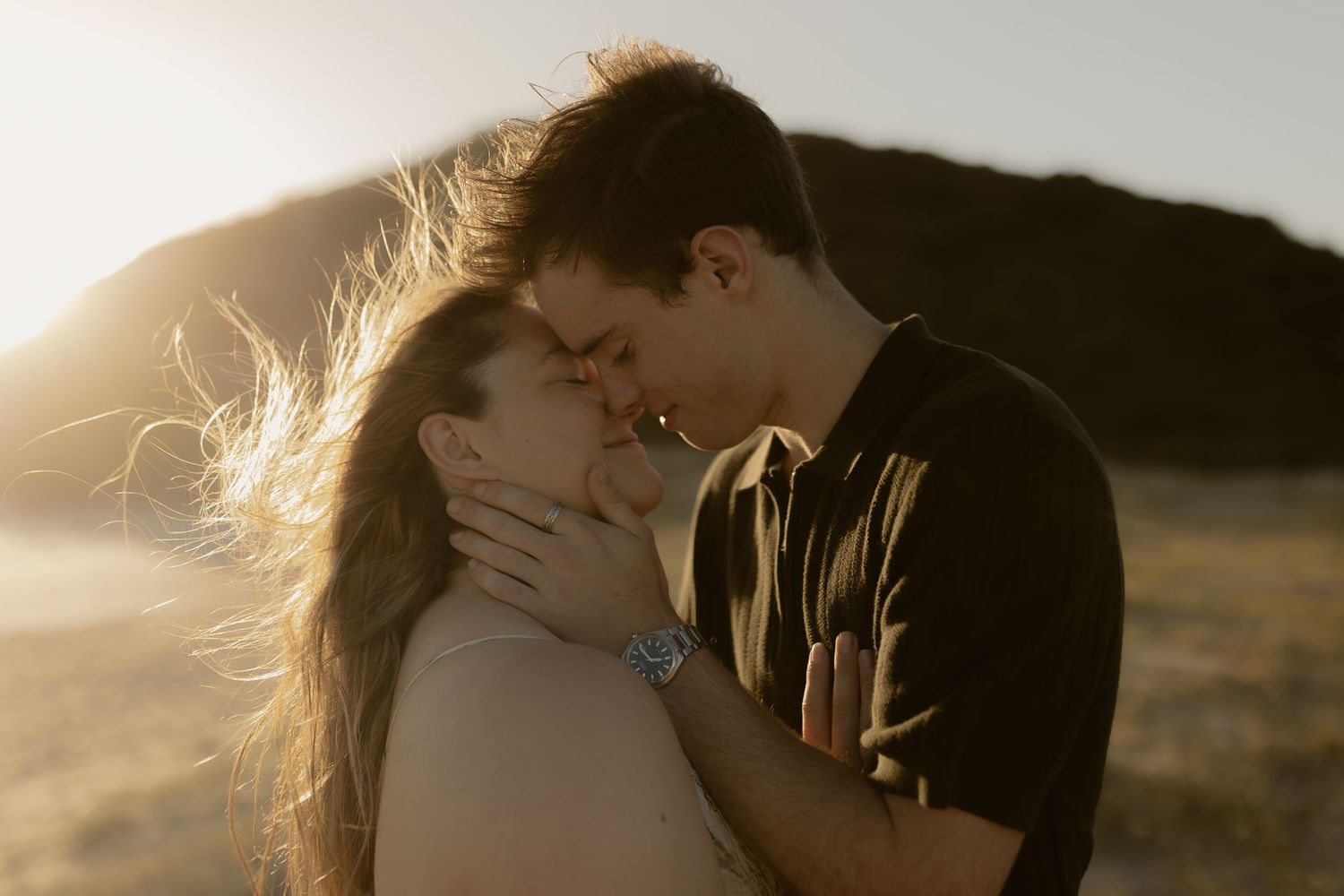 A romantic couple shares an intimate kiss at sunset with golden light and windswept hair creating a dreamy silhouette.