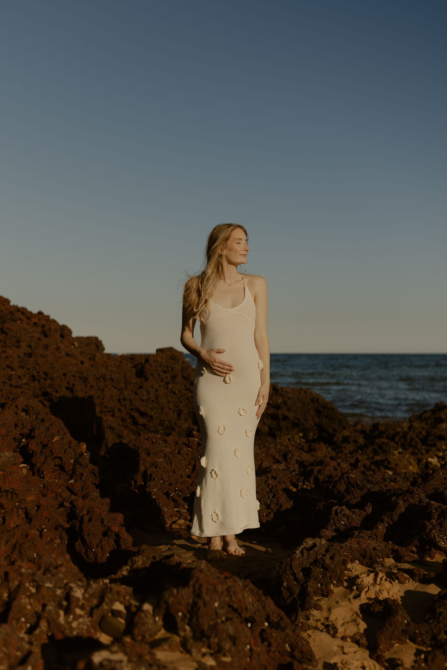 A figure in a cream-colored dress poses gracefully on rocky coastal cliffs at sunset with the ocean in the background.