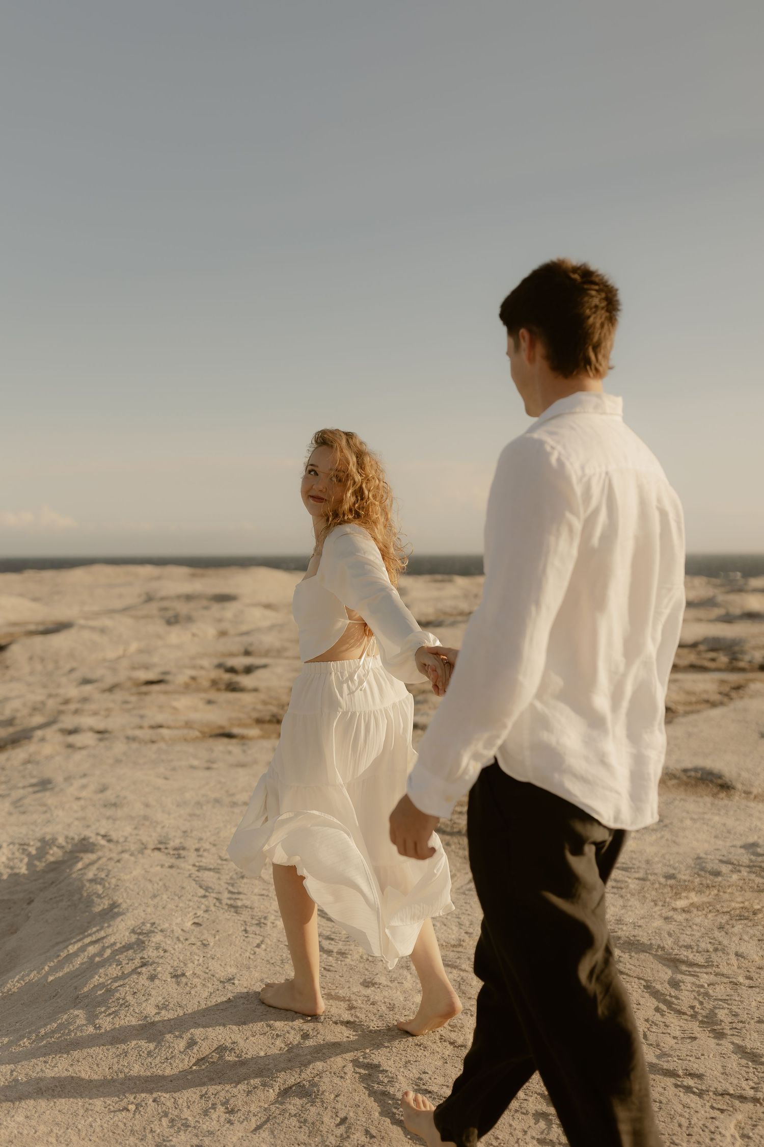 Couple in white clothing walking hand in hand on sandy beach during sunset.