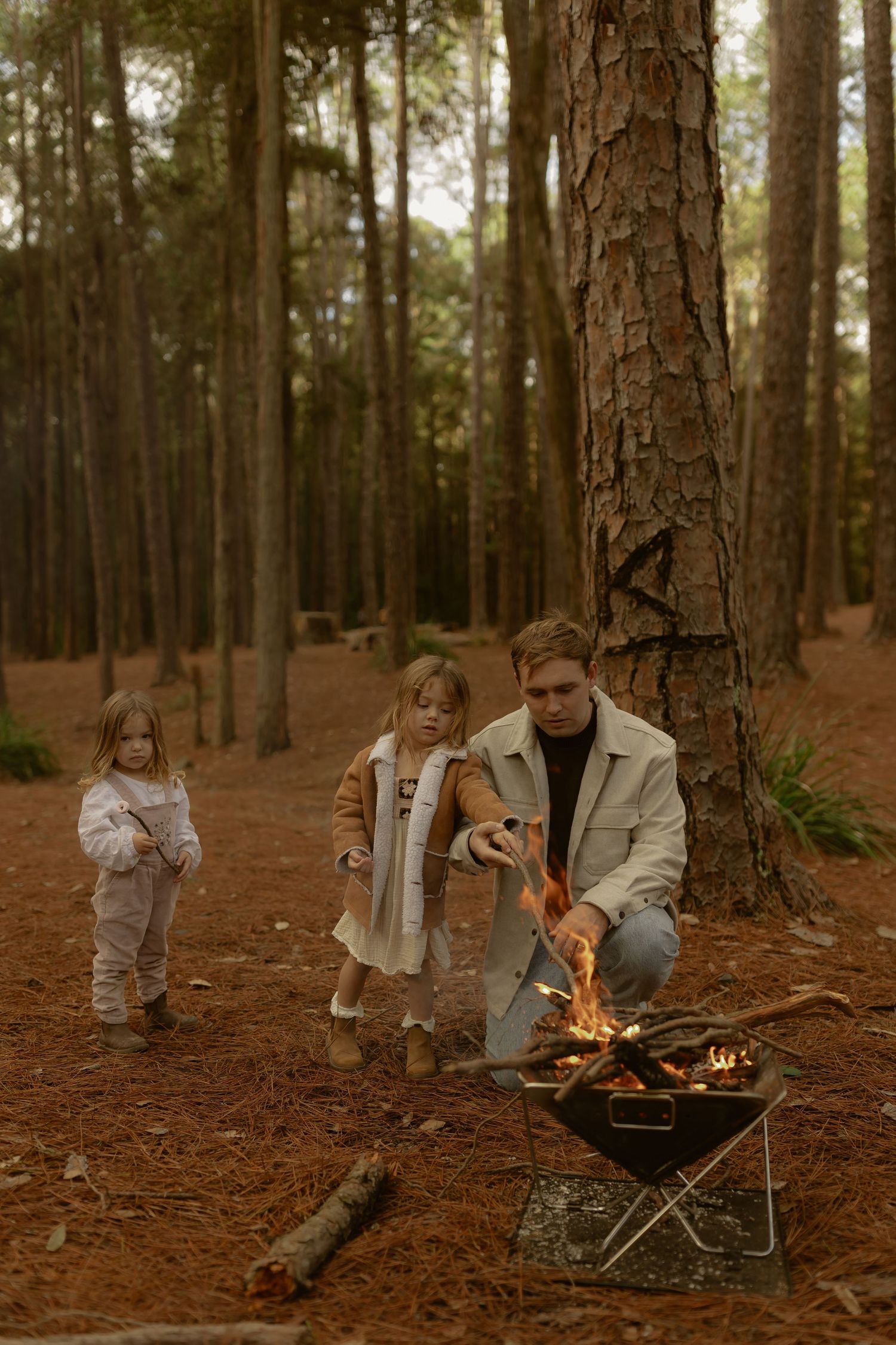 A father tends a small campfire with two young children nearby among tall pine trees in an autumn forest.