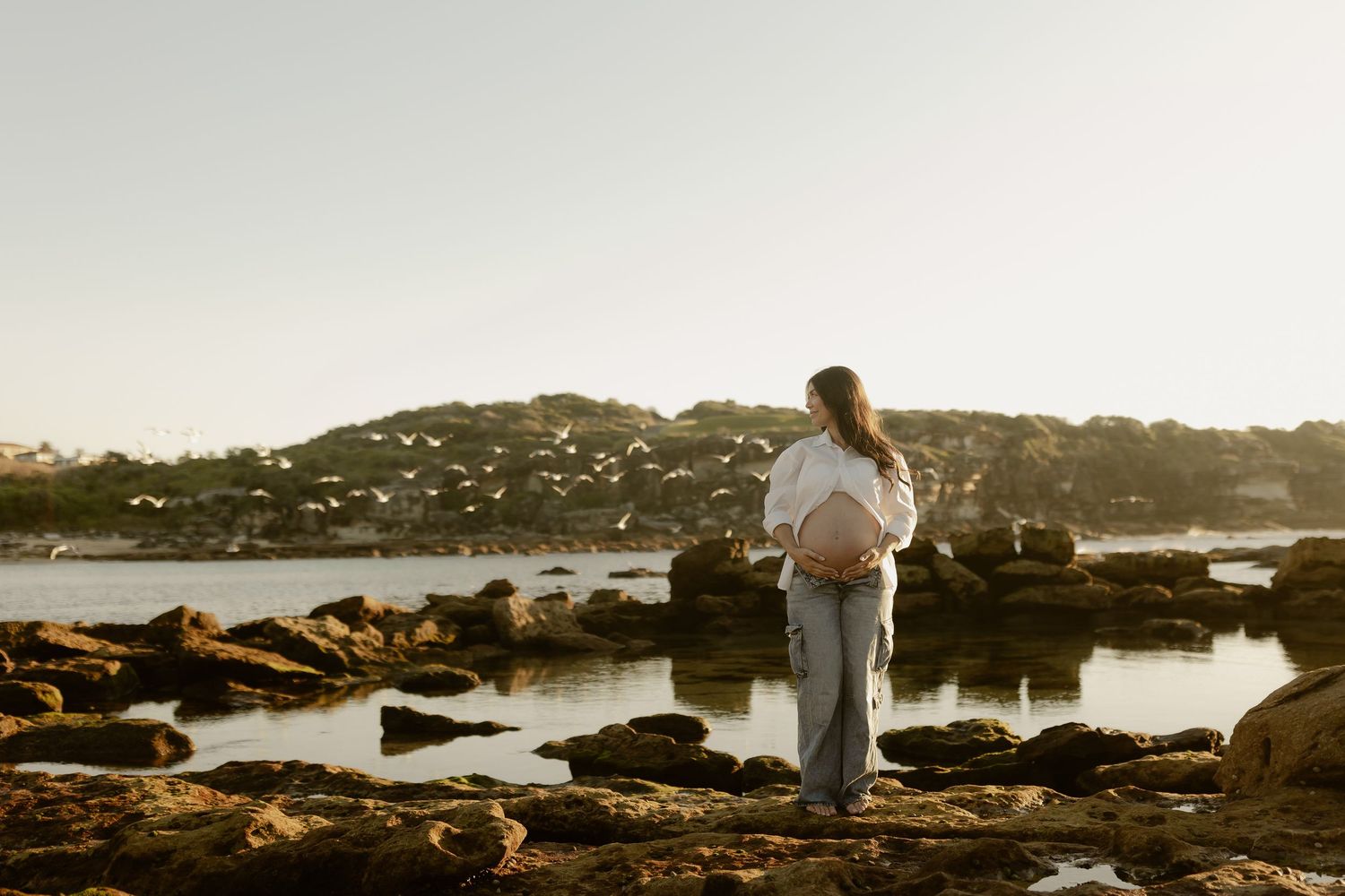 Pregnant woman standing on rocky shoreline at sunset, cradling her baby bump while gazing at a scenic coastal view.