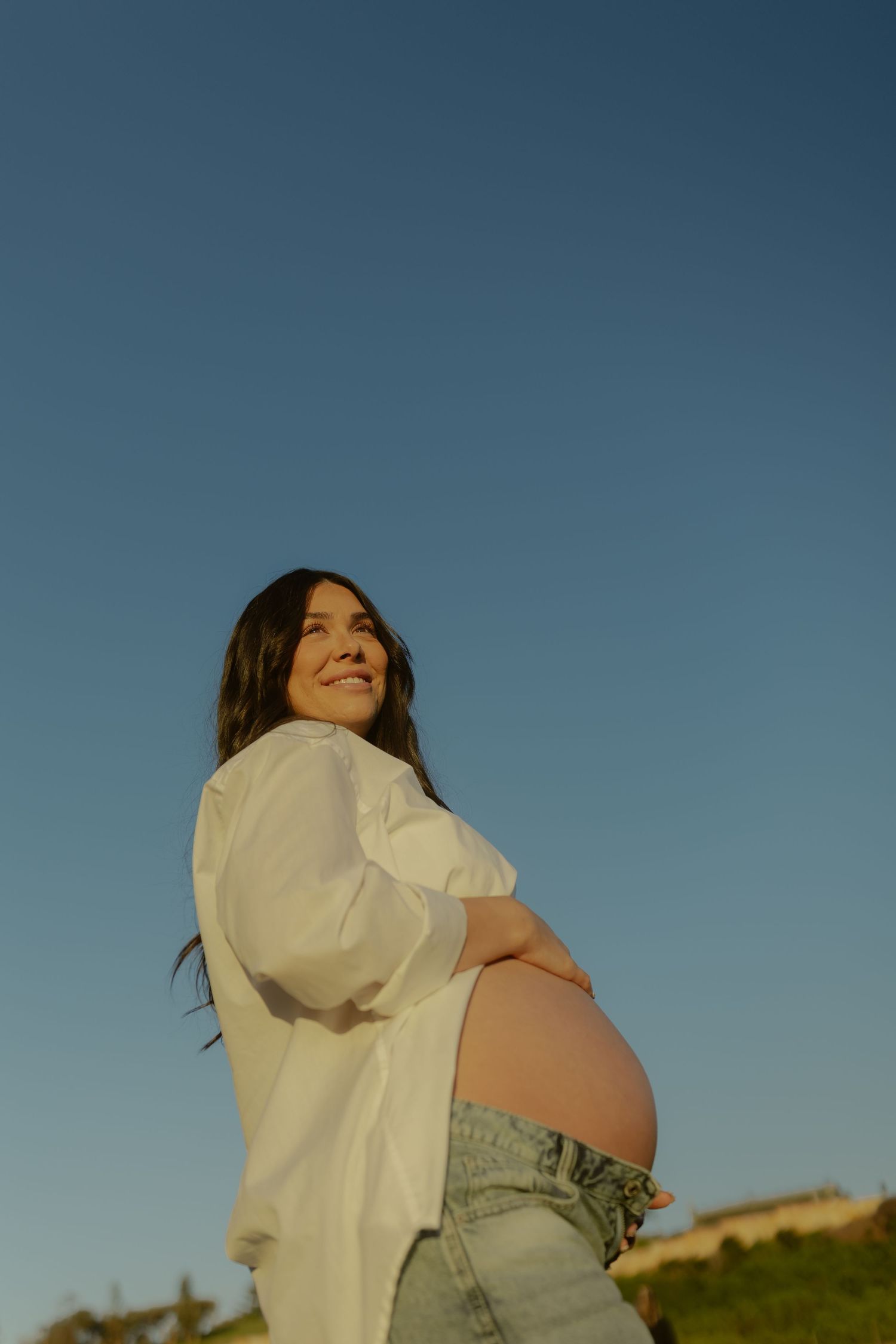 Smiling pregnant woman in white shirt holding baby bump against clear blue sky at sunset.