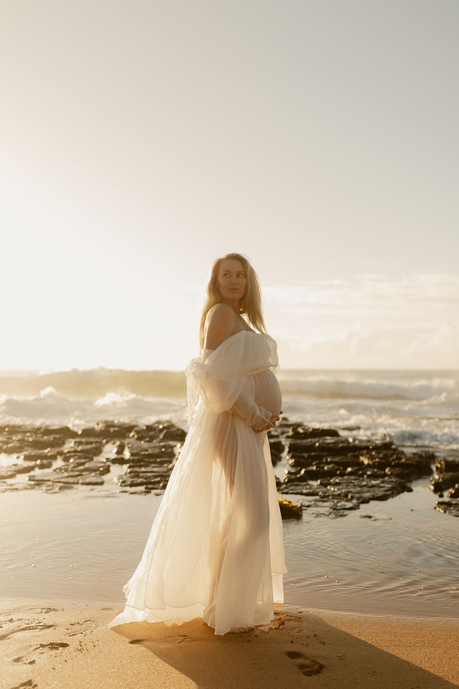 Pregnant woman in white off-shoulder gown standing on sandy beach at golden sunset with ocean waves.