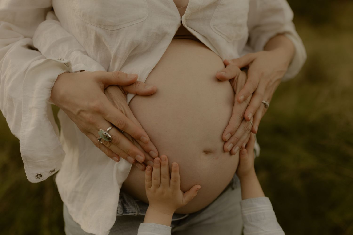 A pregnant woman in a white dress cradles her belly in a natural outdoor setting during a maternity photoshoot.