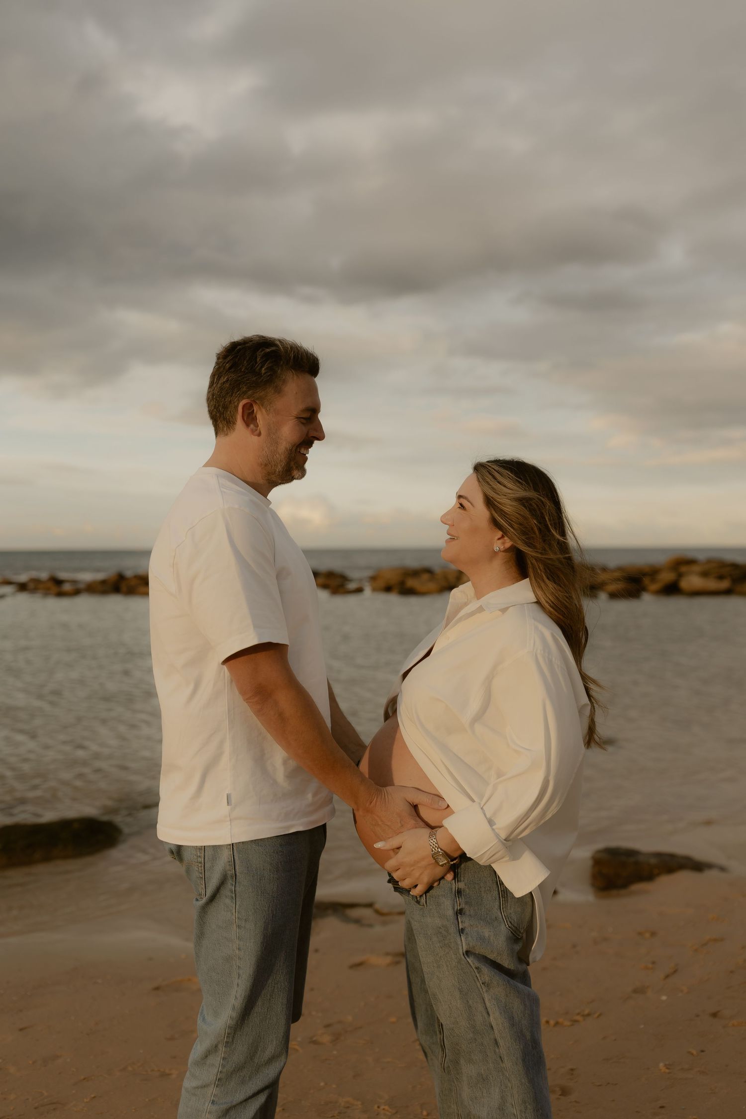 Expecting couple holds hands facing each other on a rocky beach at sunset, both wearing white tops and jeans.