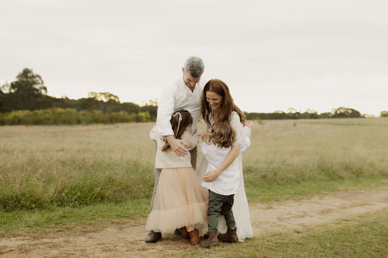 People in white clothing walk together on a winding dirt path through a sunny meadow.