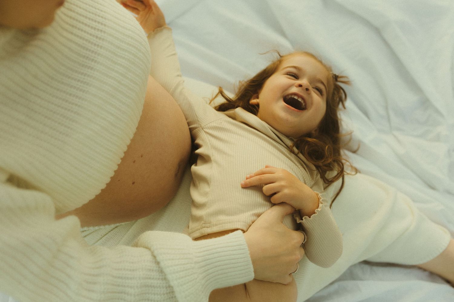 A person laughs joyfully while lying on white bedding in a cozy bedroom setting.