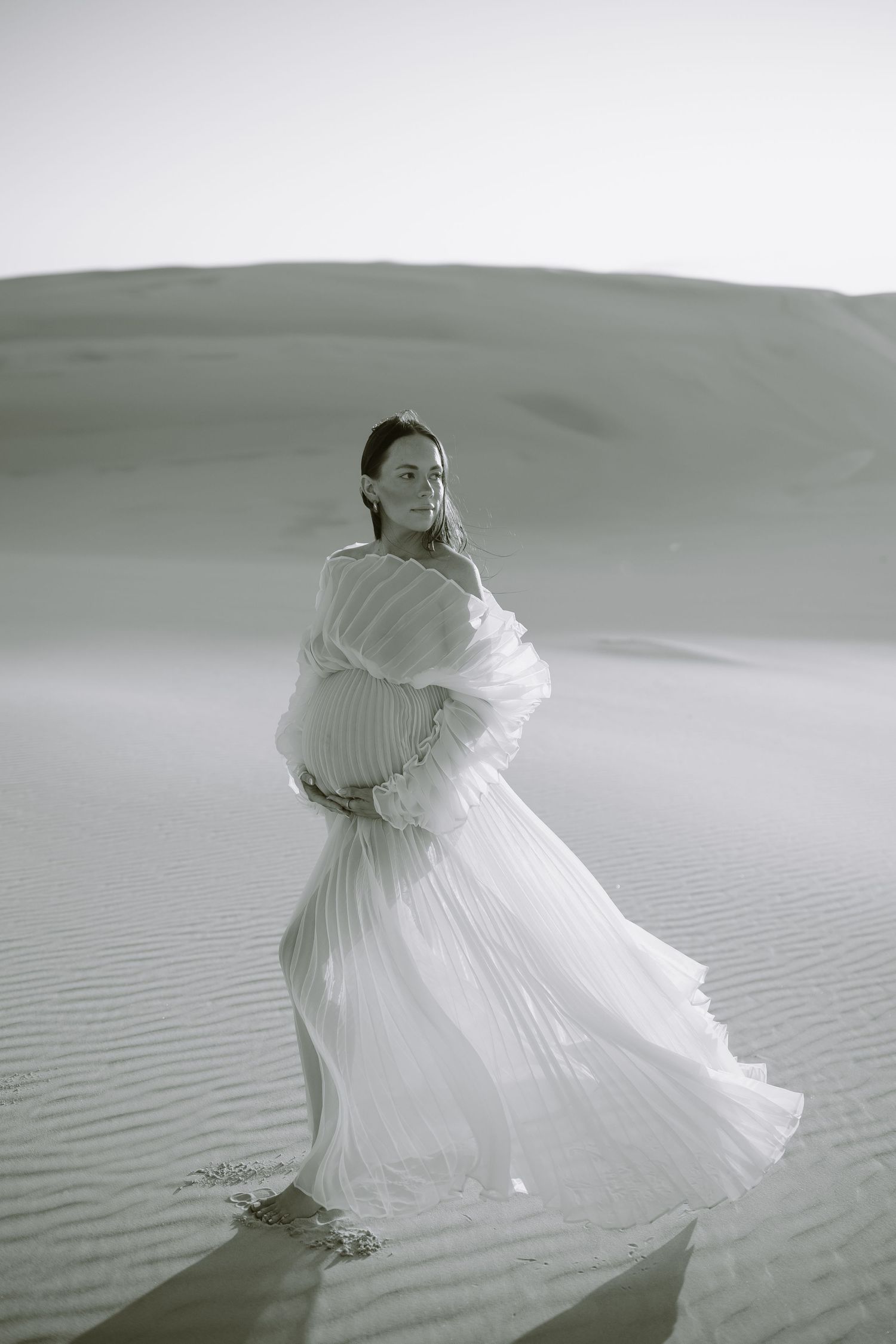 Ethereal black and white photo of a figure in flowing white dress standing against sweeping sand dunes.