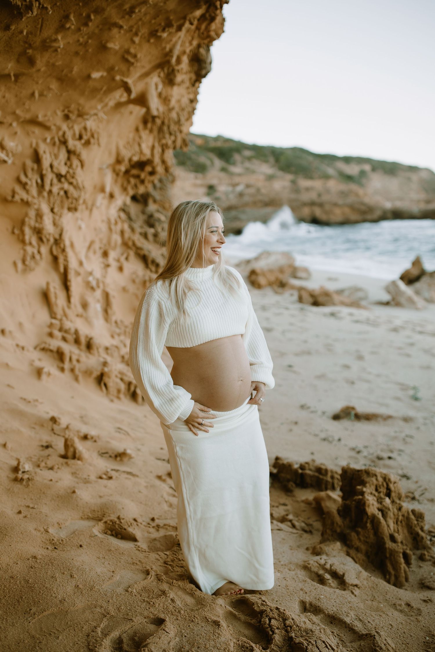 Beachside maternity photo of woman in white sweater and skirt standing against golden cliffs near ocean waves.
