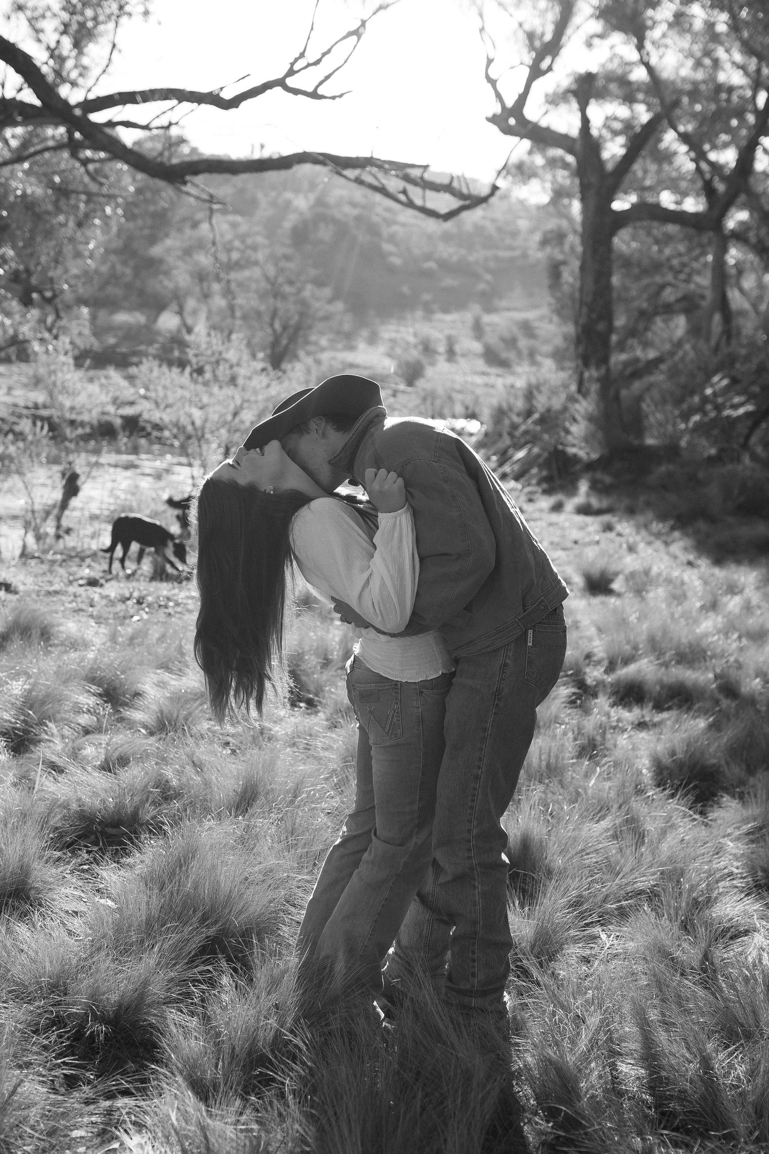 Couple embracing in black and white photo surrounded by tall grass in rustic outdoor setting.