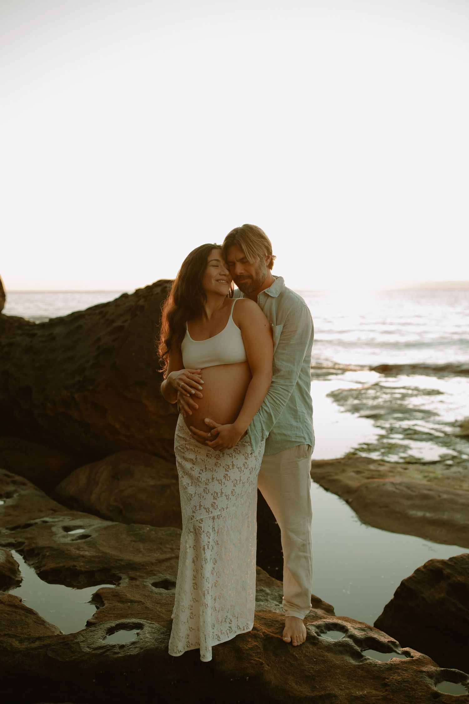 Intimate maternity photo of couple embracing on rocky beach at sunset.