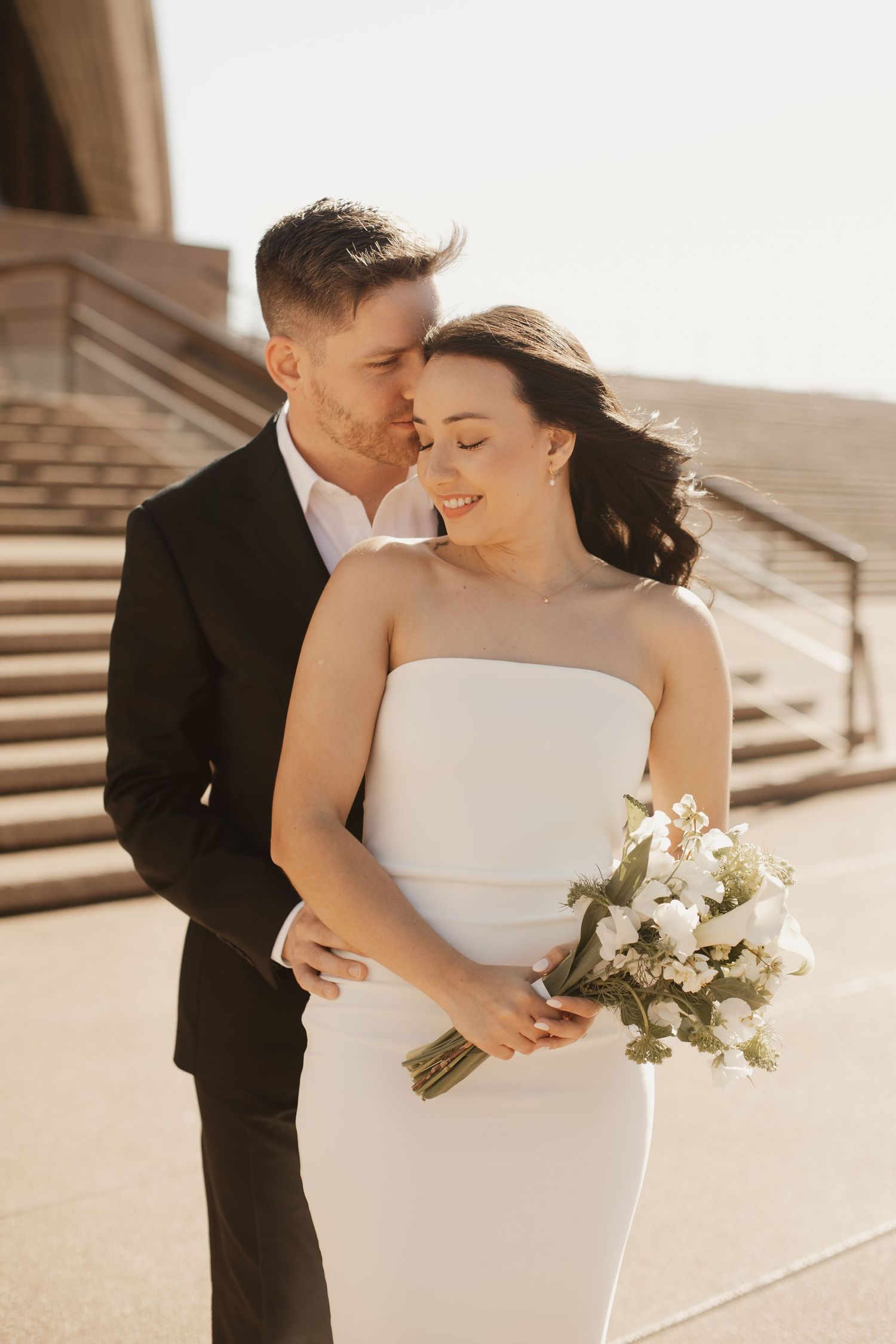 Intimate wedding photo with couple embracing on stone steps at sunset, bride holding white bouquet in sleek dress.