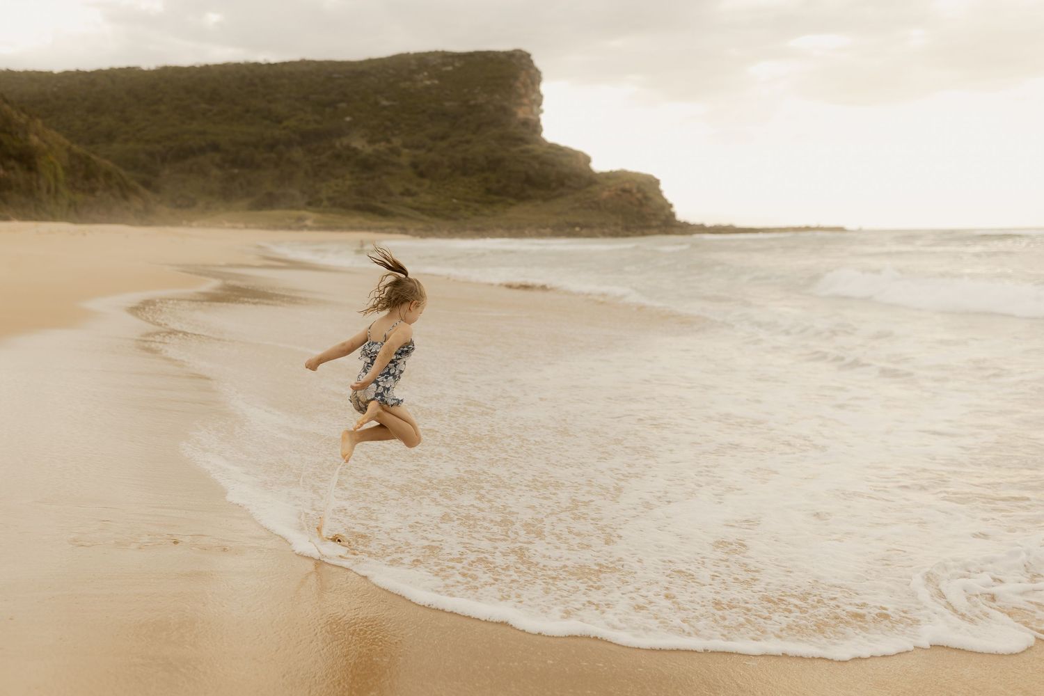 A figure leaps joyfully along a sandy beach at sunset with waves crashing in the background.