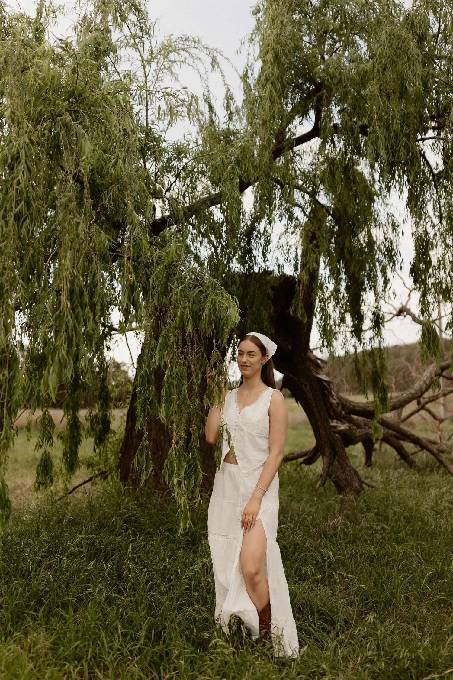 A person in a flowing white dress poses beneath a dramatic weeping willow tree.