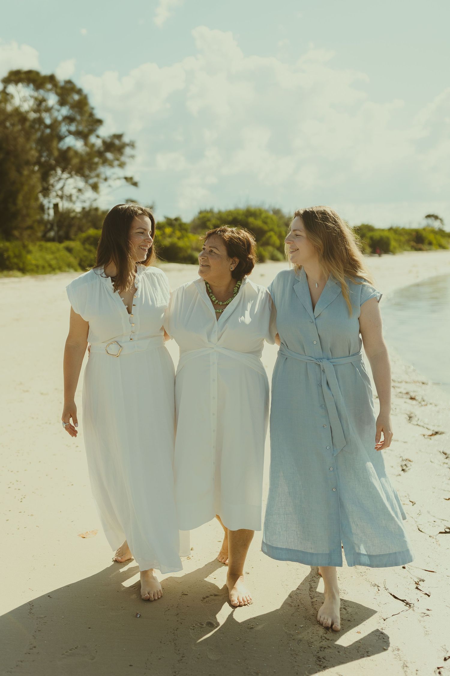 Three people in coordinating white and blue outfits stroll barefoot along a pristine sandy beach.