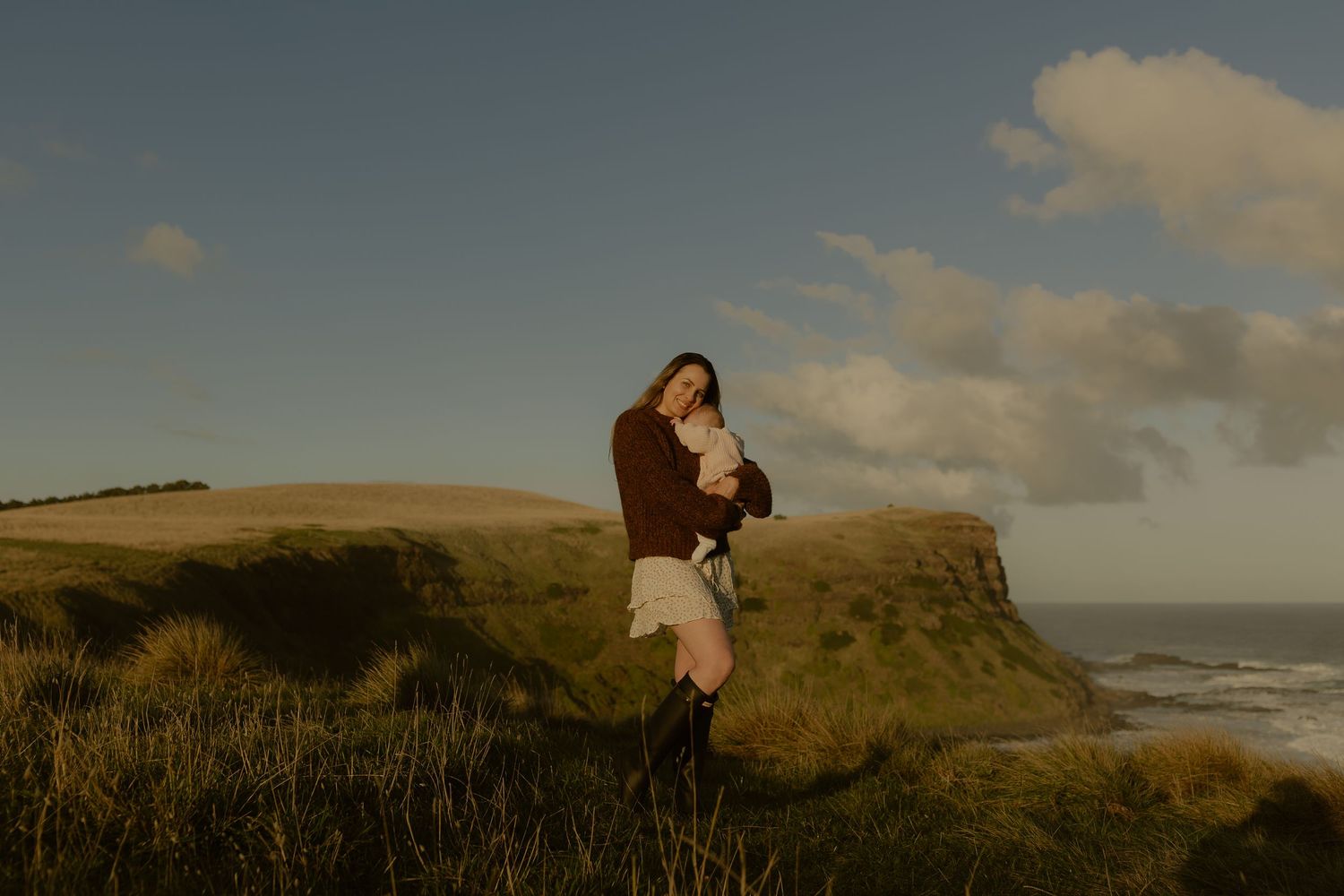 Woman holding baby on scenic coastal hillside with ocean cliffs and dramatic sky at golden hour.