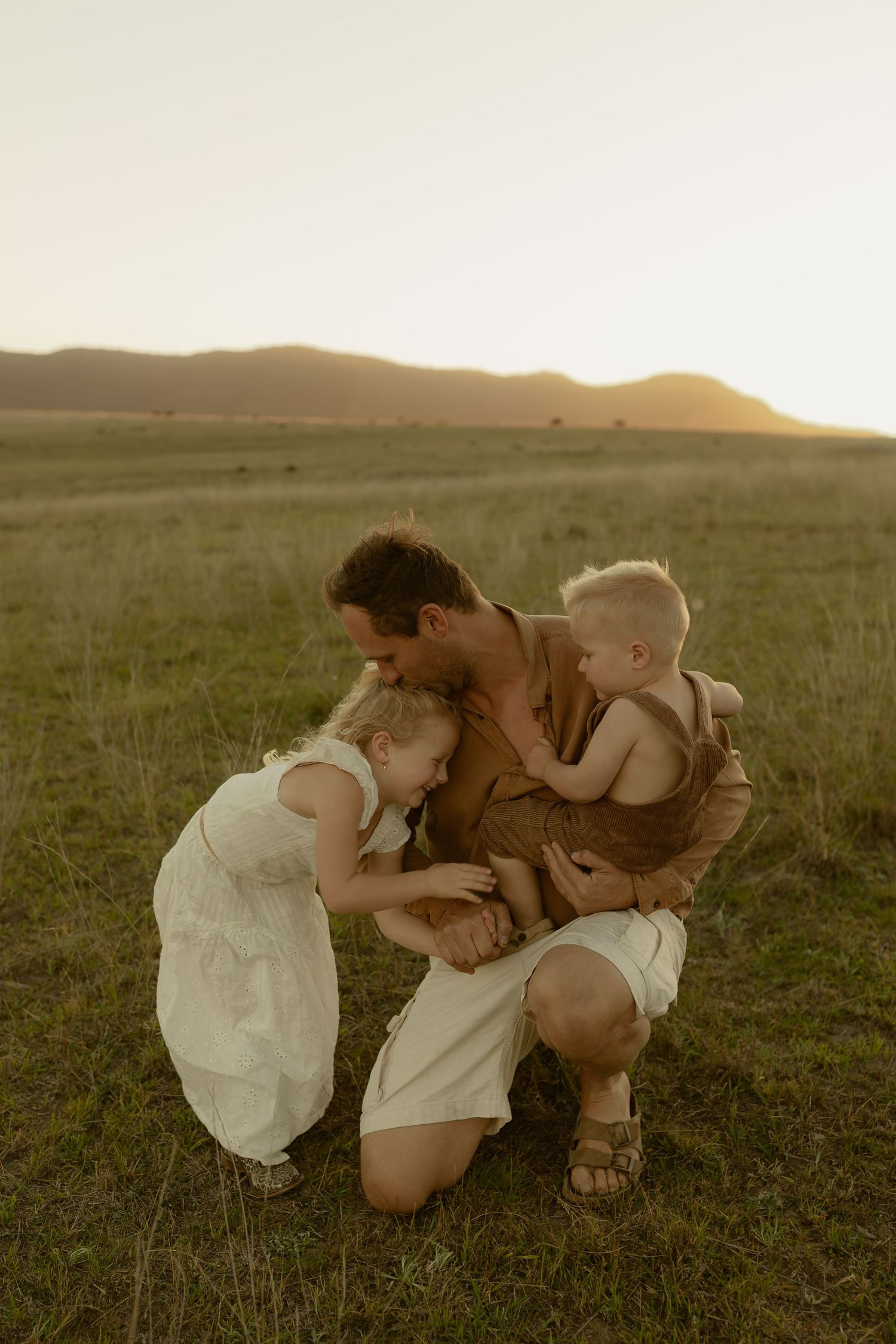 A parent kneels in a golden field at sunset, tenderly holding two young children in a heartwarming family moment.