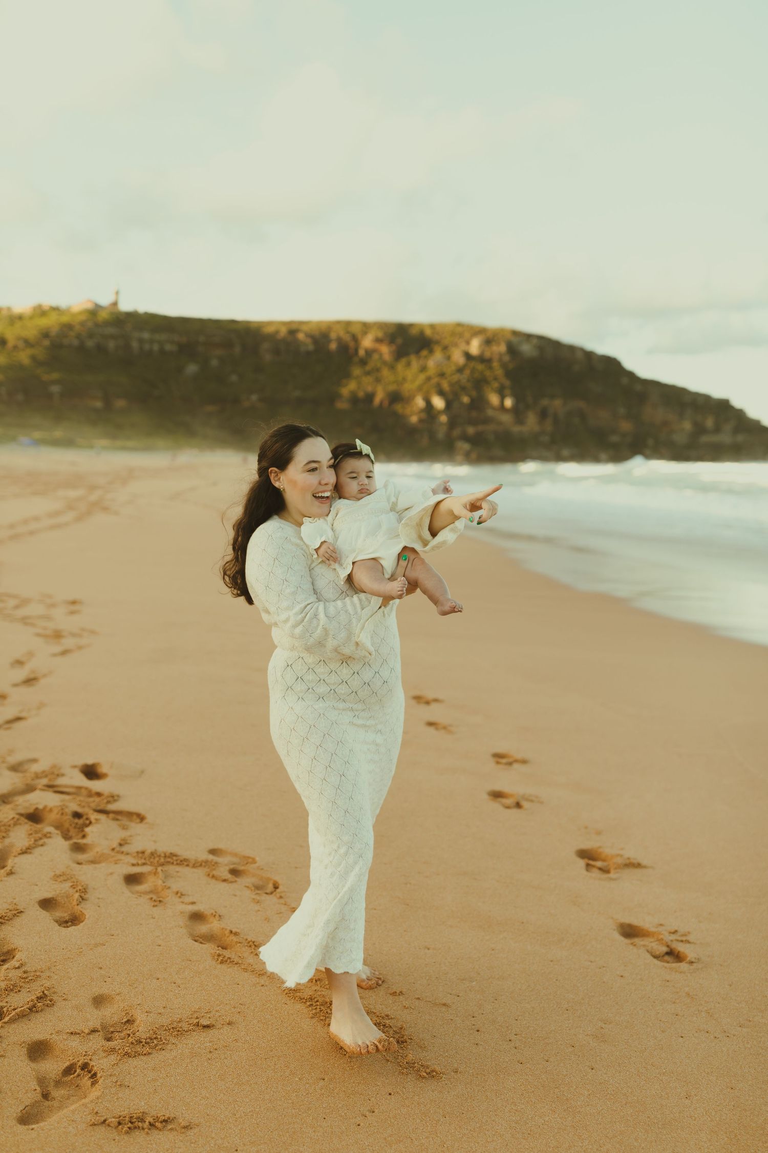 Two people in white dresses dance joyfully together on a sandy beach at sunset with ocean waves in the background.