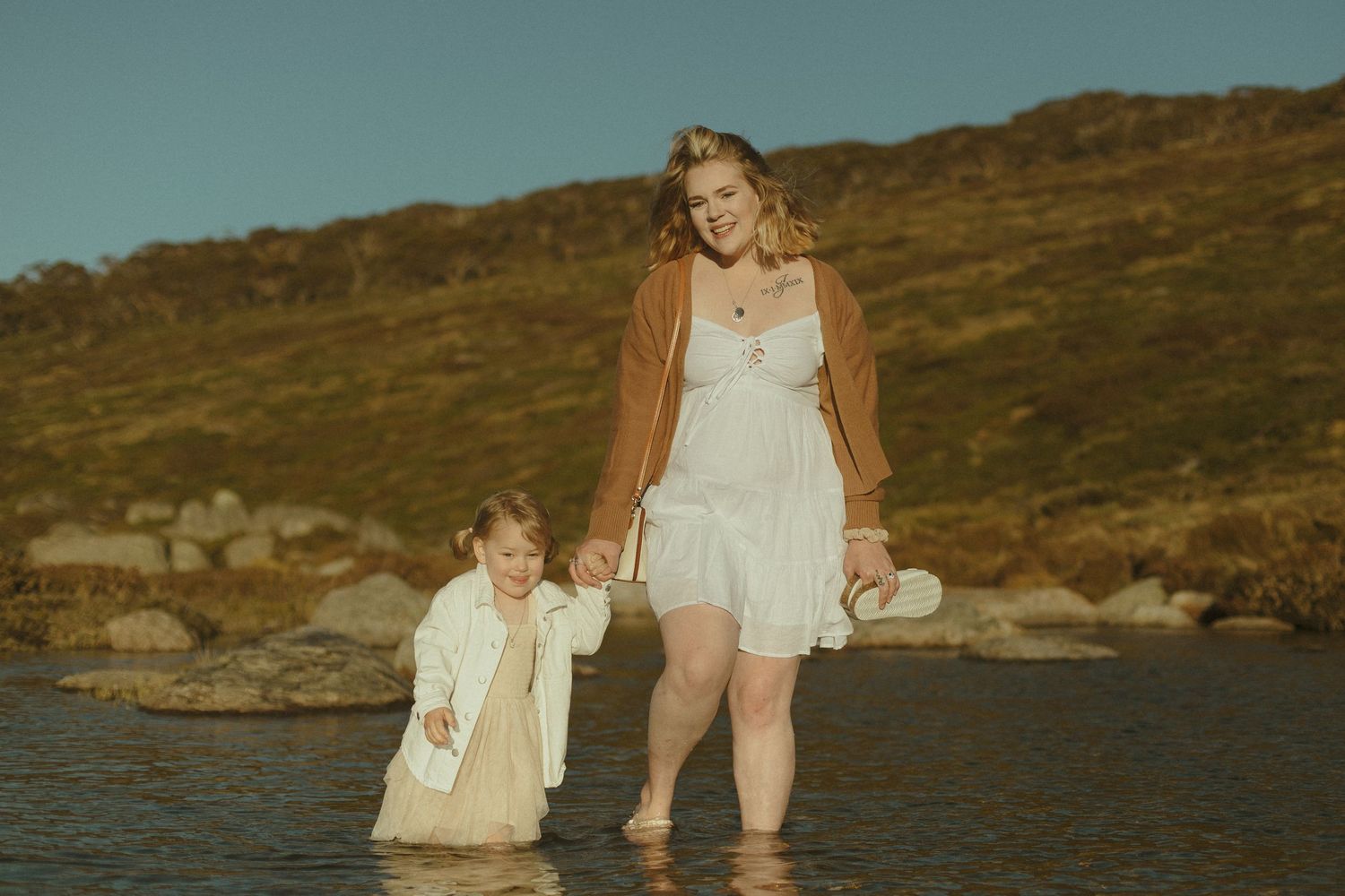 Two people wearing white outfits standing in shallow water at sunset with hills in the background.