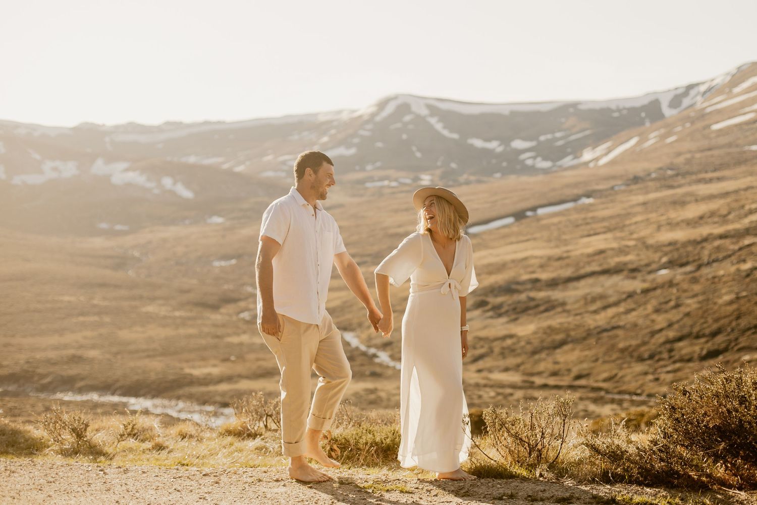 A couple in light colored clothing stands on a mountain trail at sunset with rolling hills in the background.