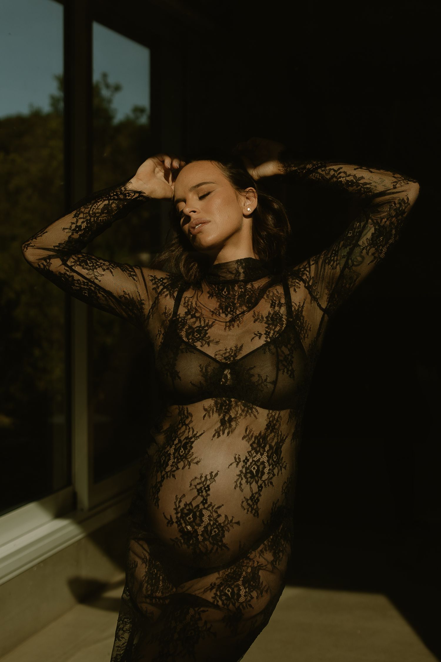 Silhouetted figure in black lace maternity dress poses by window in warm golden sunlight.