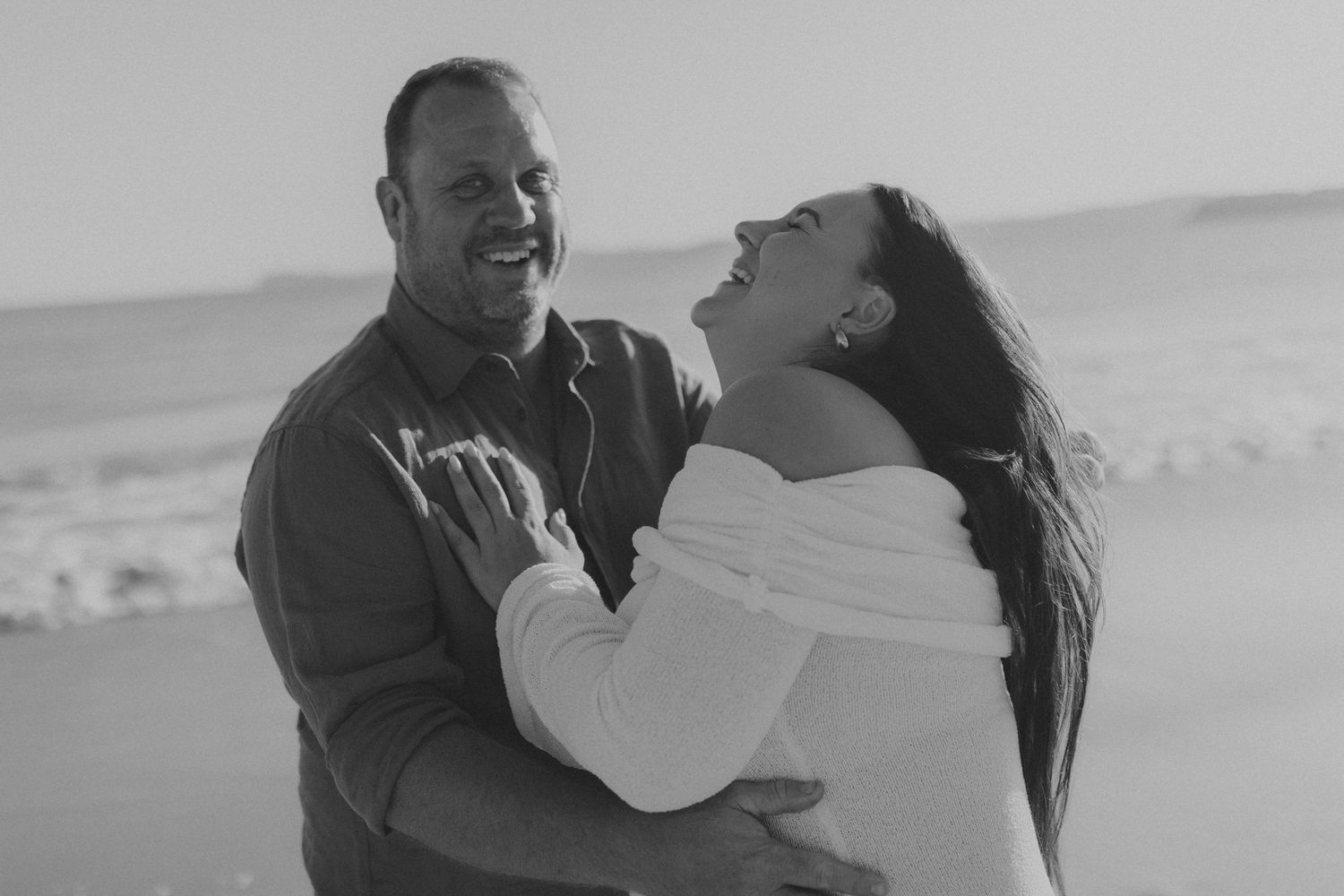 A black and white photo series showing a couple embracing and laughing together on a beach.