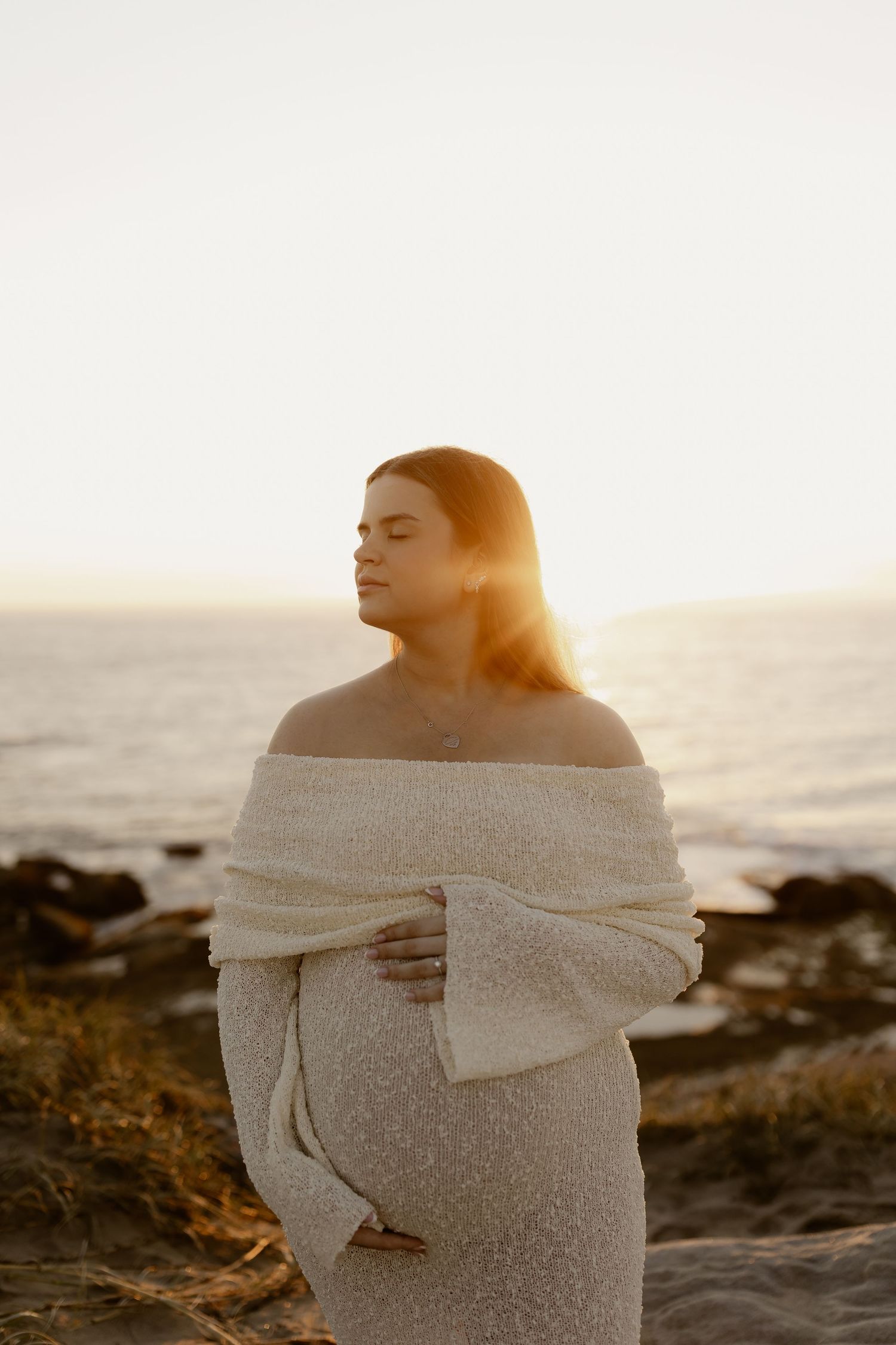 A person wrapped in a white blanket stands on a rocky beach at sunset with warm golden light illuminating the scene.
