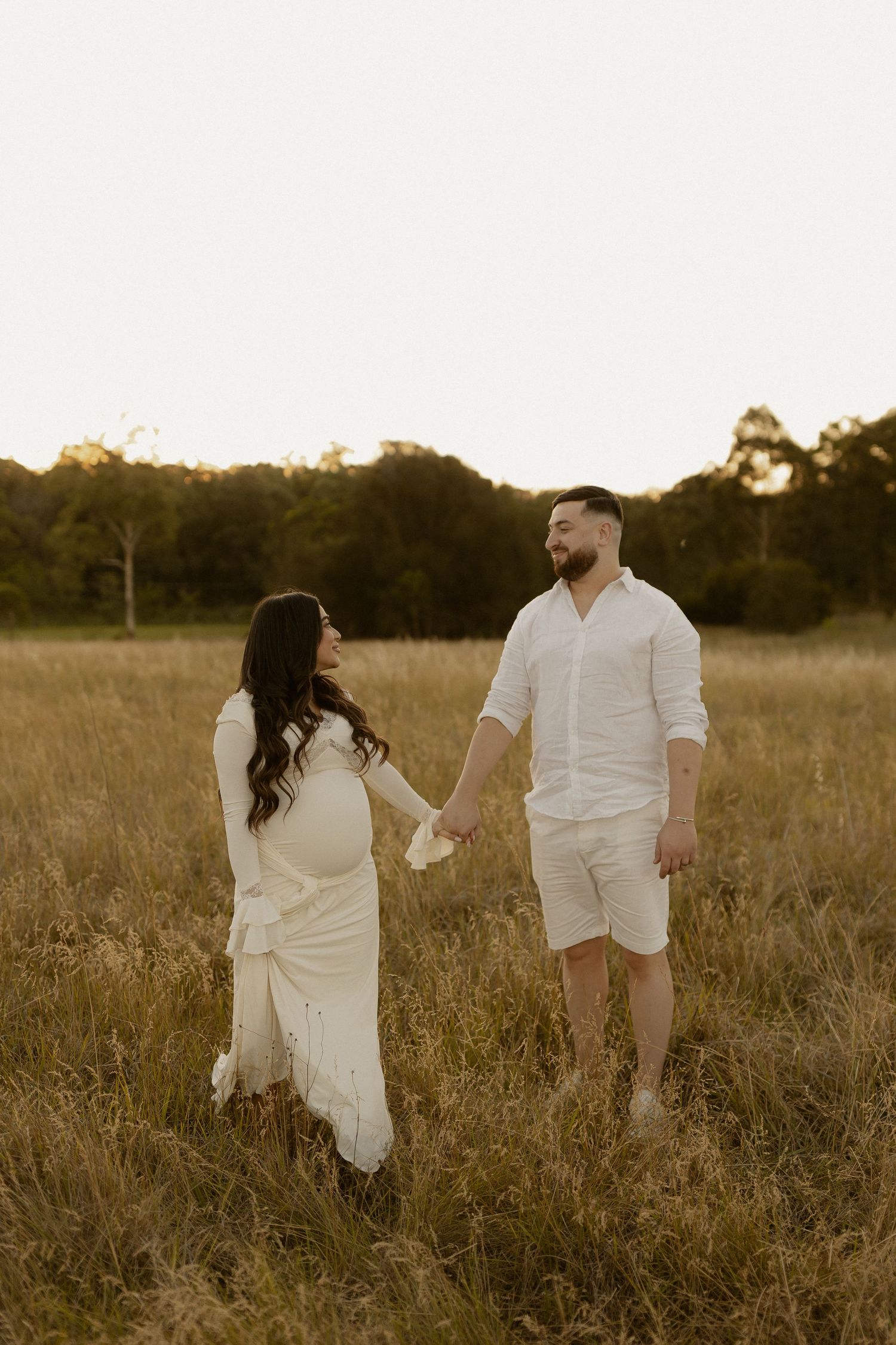 Expecting couple holding hands while walking through tall grass at golden hour.