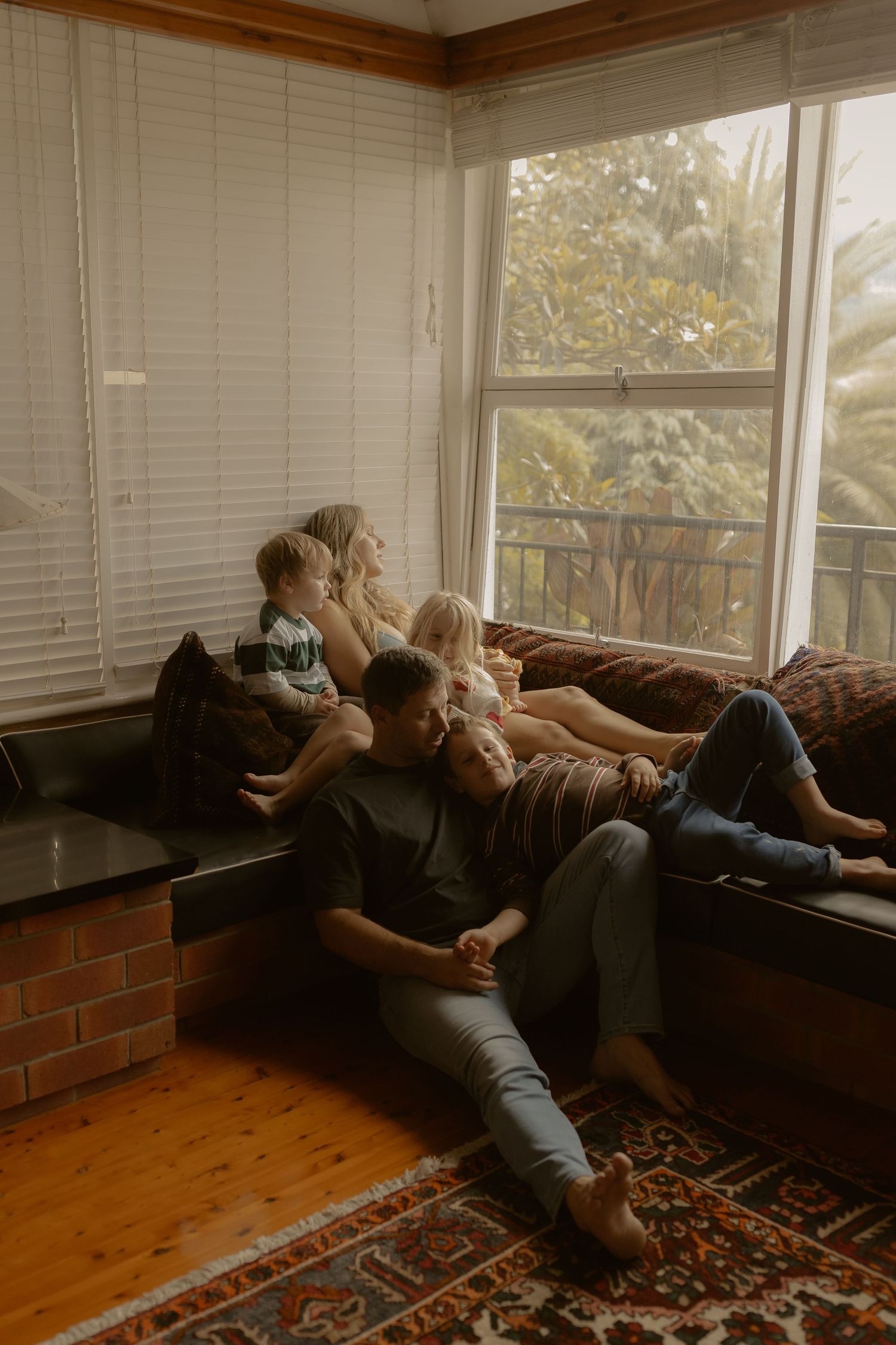 A family relaxes together on a window seat sofa, with soft natural light filtering through white blinds indoors.
