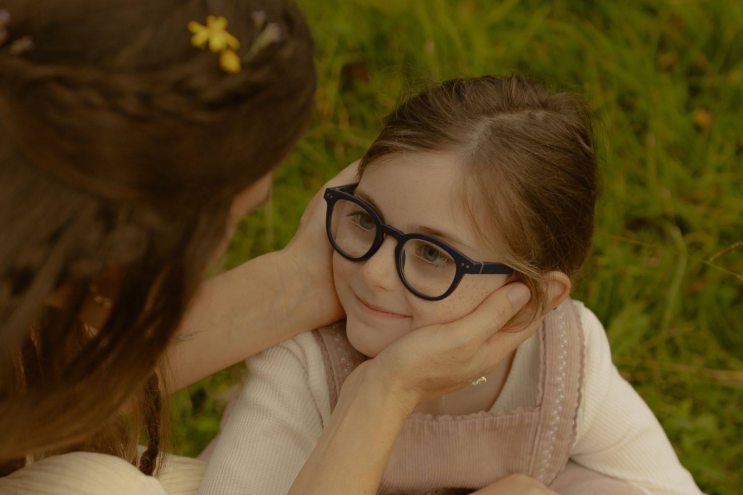 A young girl with glasses rests her chin on her hands while an older girl gently cups her face outdoors.
