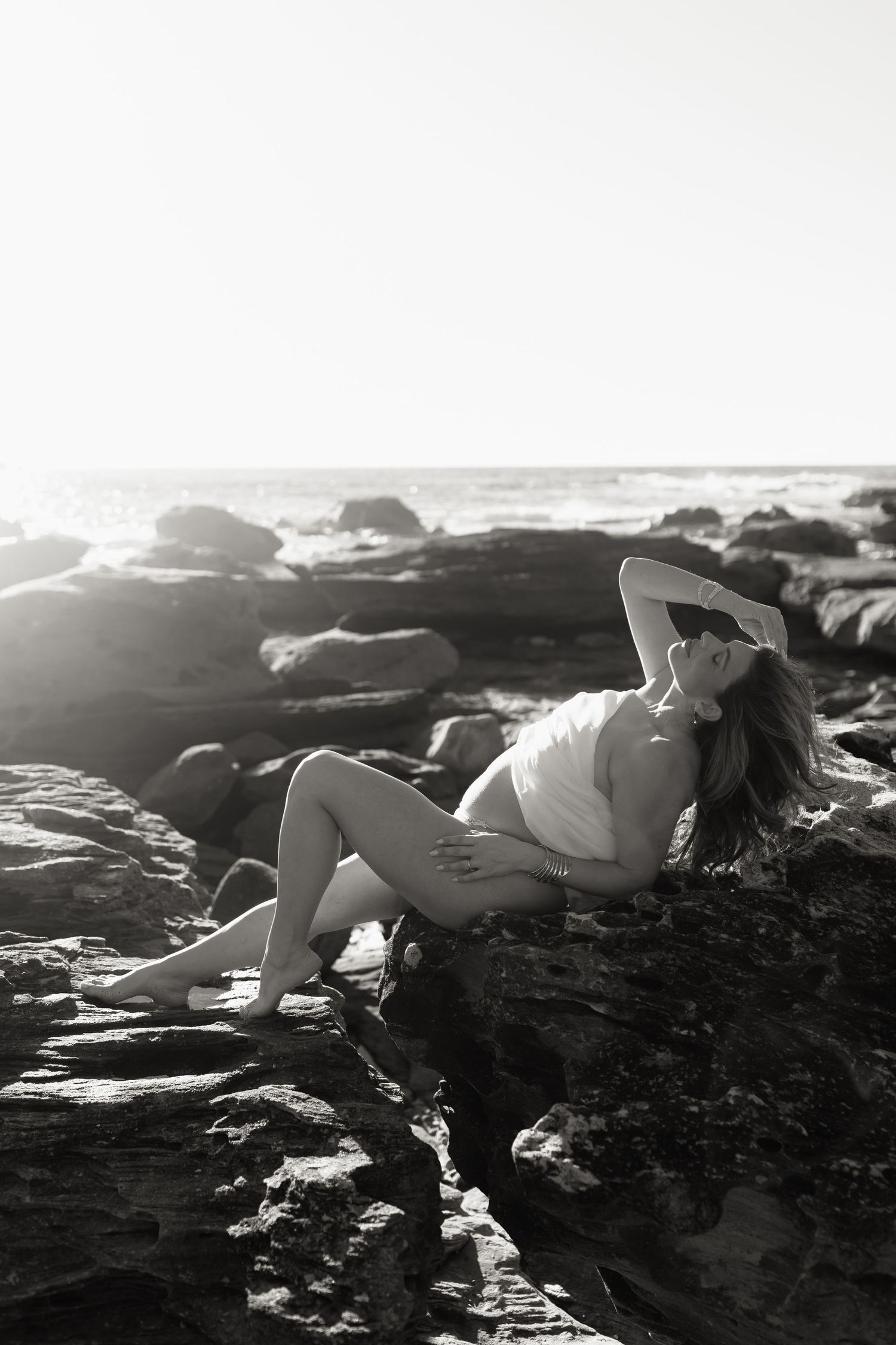 Black and white photo of pregnant woman reclining on coastal rocks with ocean backdrop in bright sunlight.