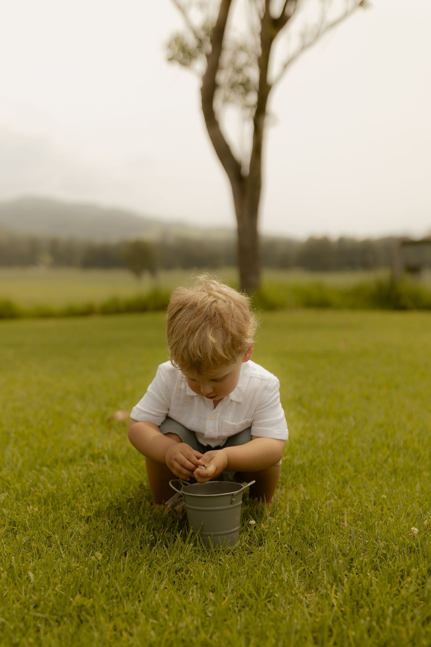 A small child crouches in a grassy field examining a metal bucket at sunset.