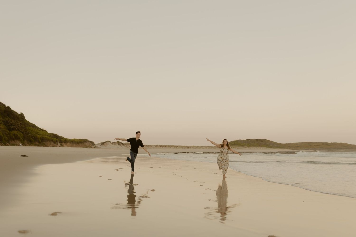 Two silhouetted figures walk along a misty beach at sunset with mountains in the distance.