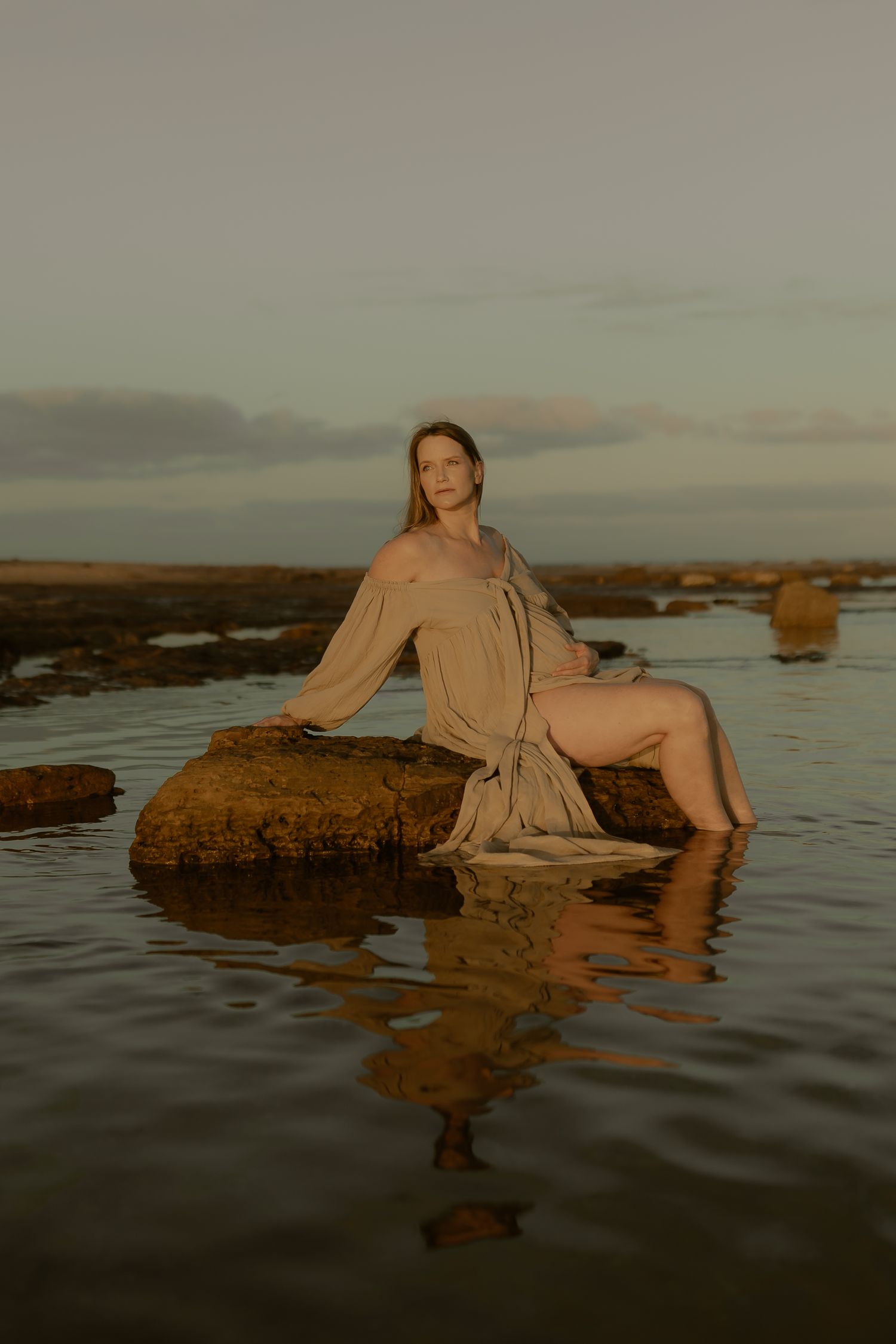 A person in a beige dress poses artistically on rocks in shallow coastal waters during golden hour.