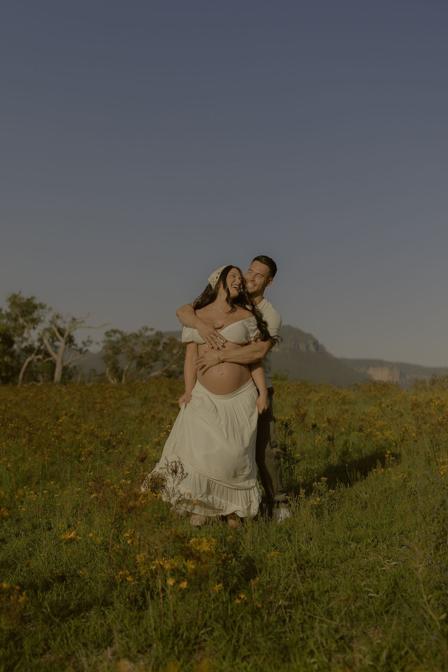 A romantic couple embraces in a golden meadow at sunset with mountains in the distance.