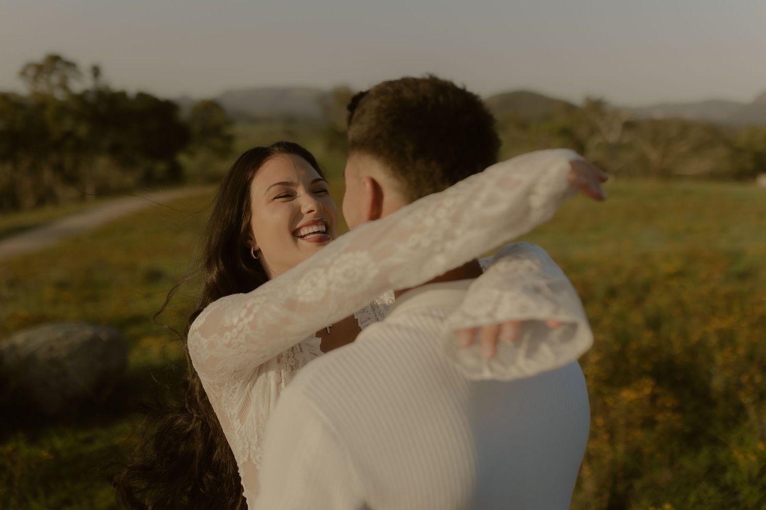 A couple embraces and laughs together in a field during sunset with mountains visible in the background.