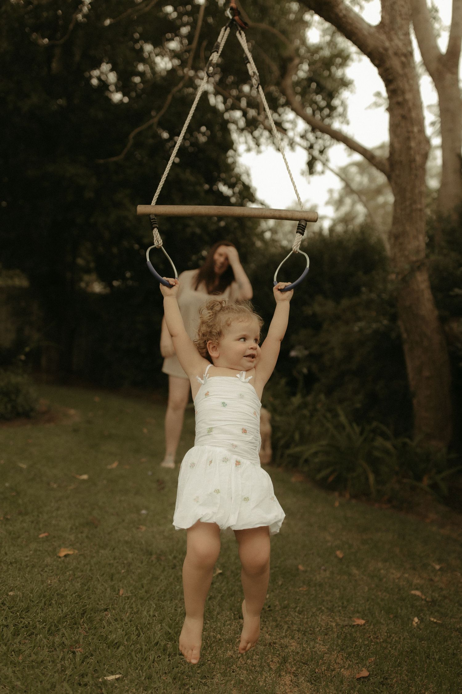 A toddler in a white dress plays on a wooden swing set in a backyard setting.