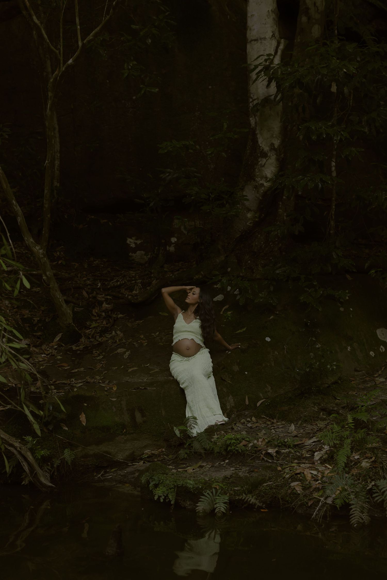 Pregnant woman in white crop top and skirt poses by forest creek surrounded by lush dark greenery.