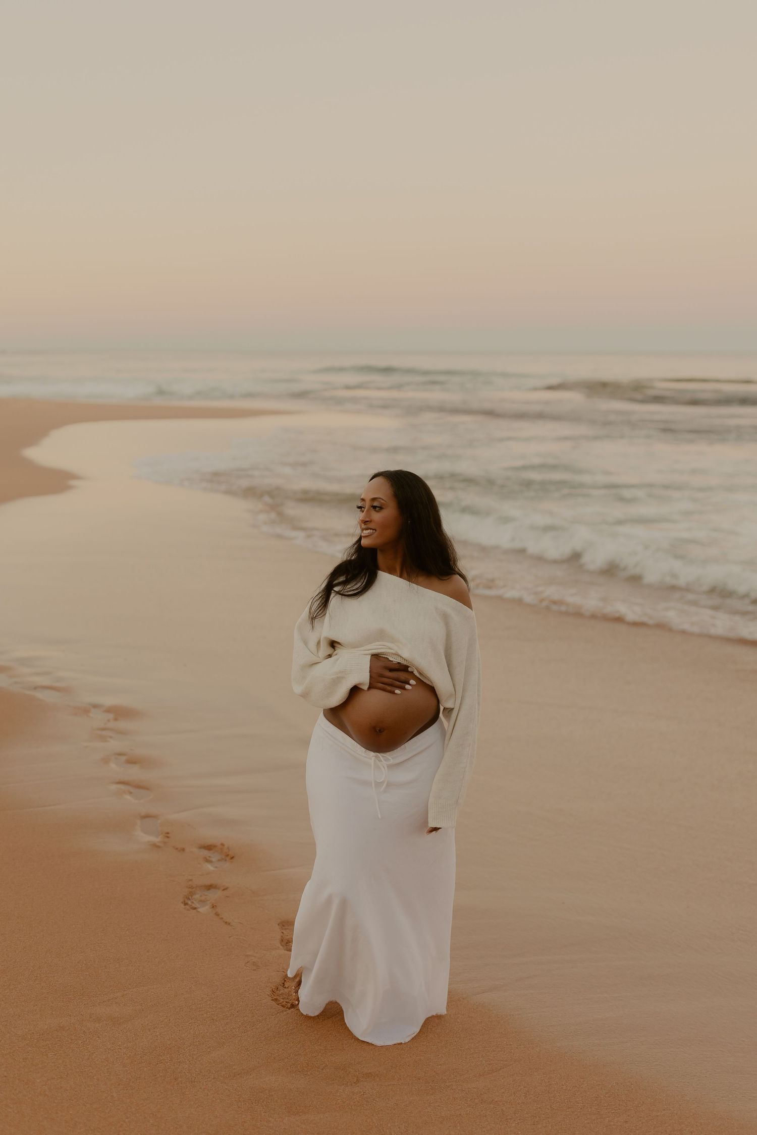 Pregnant woman in white off-shoulder top and skirt cradling baby bump on beach at sunset.