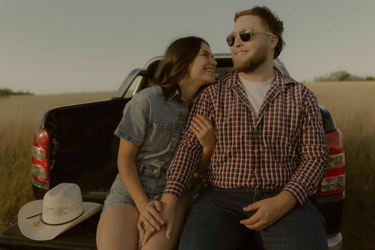 Couple sitting on truck bed in open field, woman in denim shirt leans on man in plaid shirt wearing sunglasses at dusk.