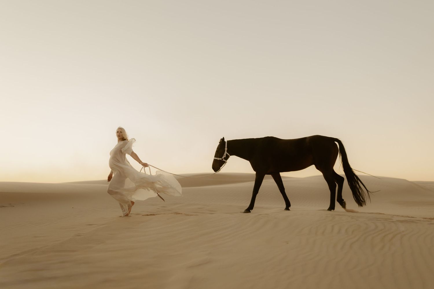 Woman in white dress leading a dark horse across golden desert sand dunes in soft warm light.