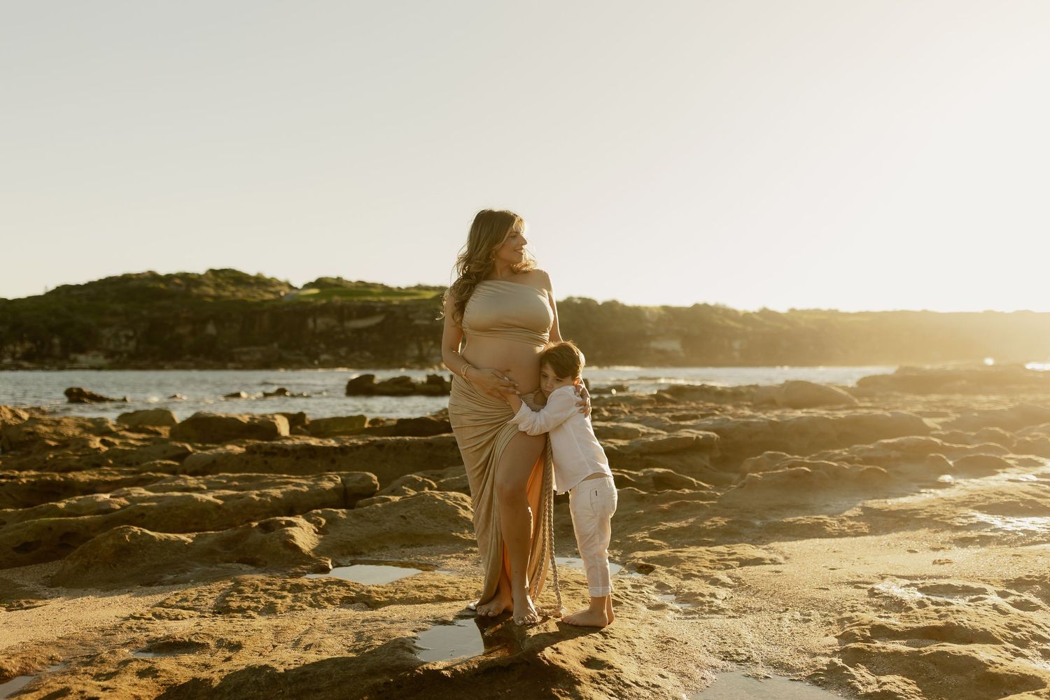 Pregnant woman standing on rocky beach at sunset with young child hugging her belly in golden light.