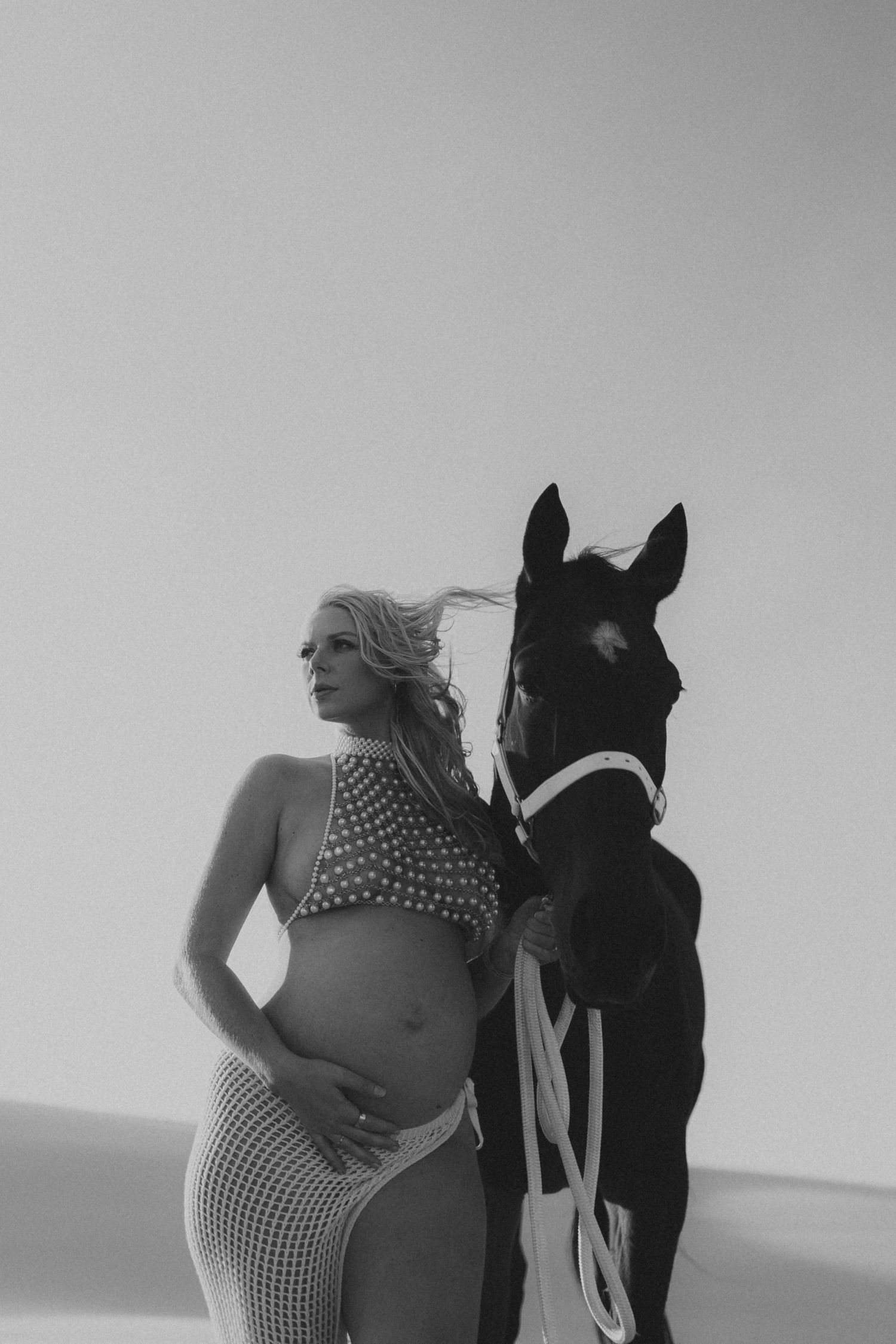 Black and white maternity photo of pregnant woman in crop top and mesh skirt posing beside a dark horse outdoors.