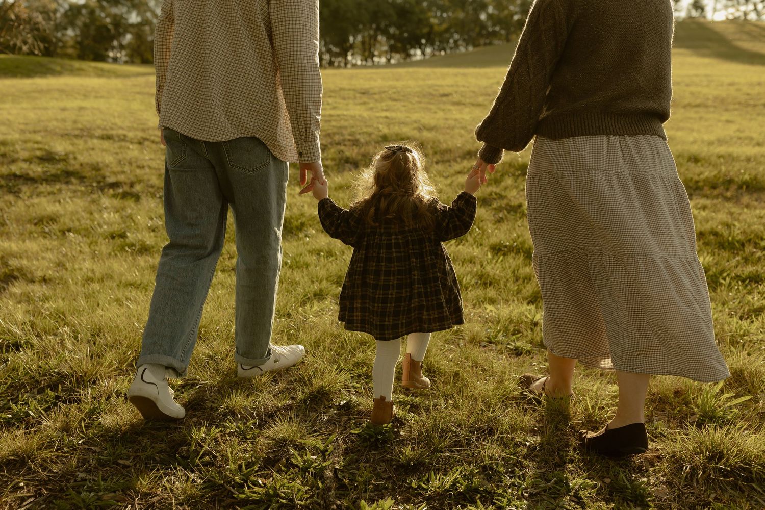 Family of three holding hands walking through a sunny green field, parents swinging young child between them.