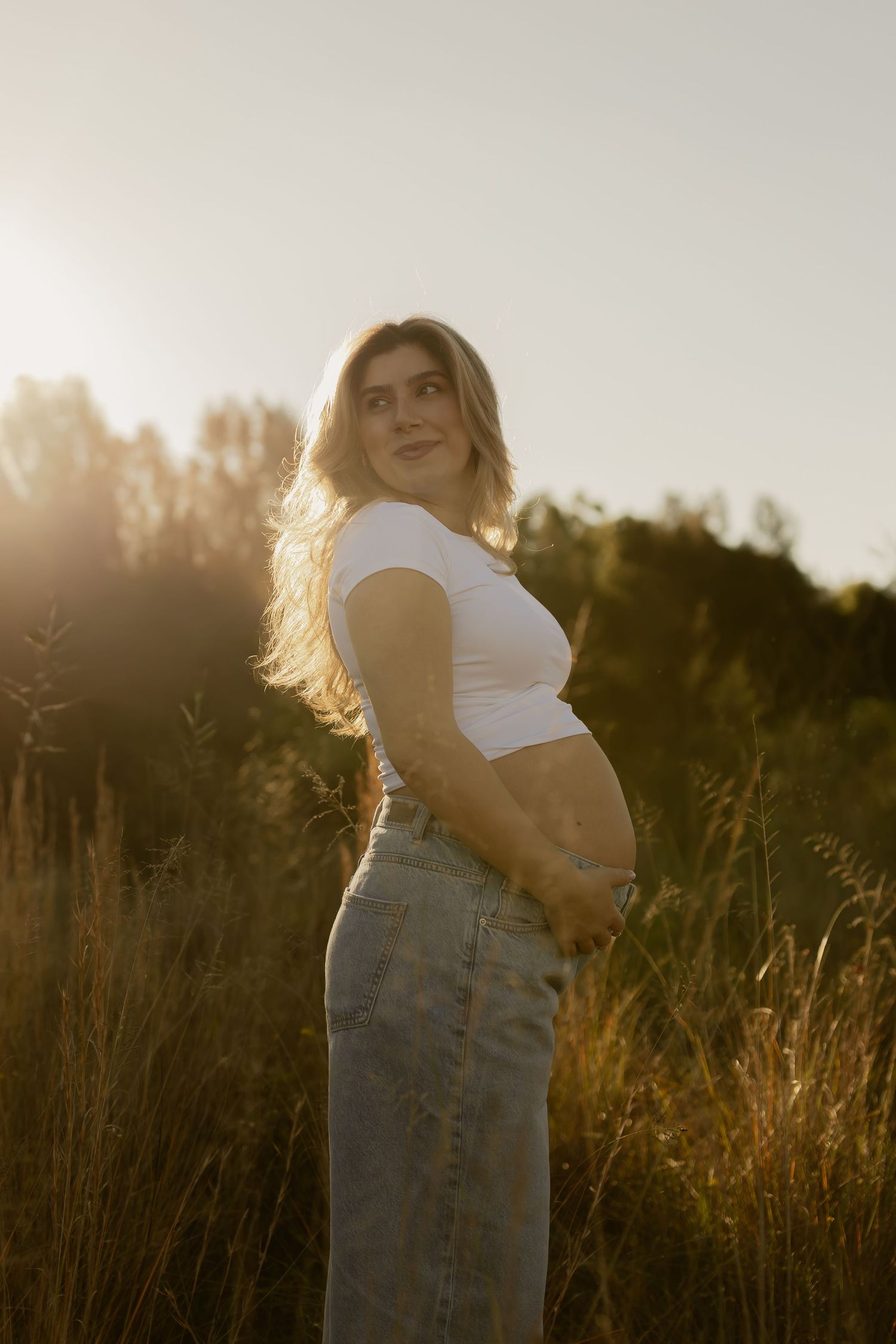 Pregnant woman in white crop top and jeans standing in golden sunlit field, cradling baby bump at sunset.