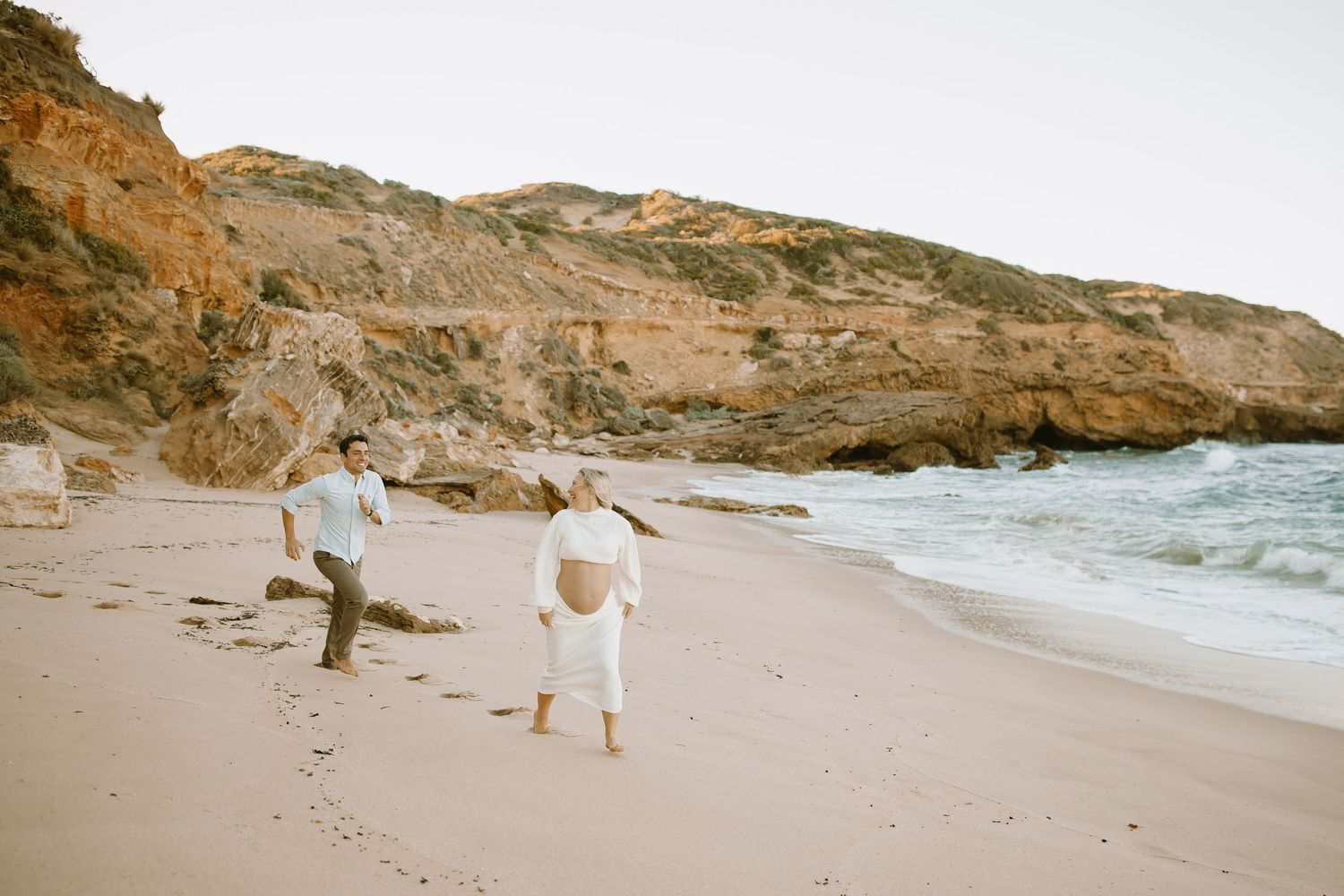 Two people walking along a scenic rocky beach coastline with waves crashing on the shore on a cloudy day.