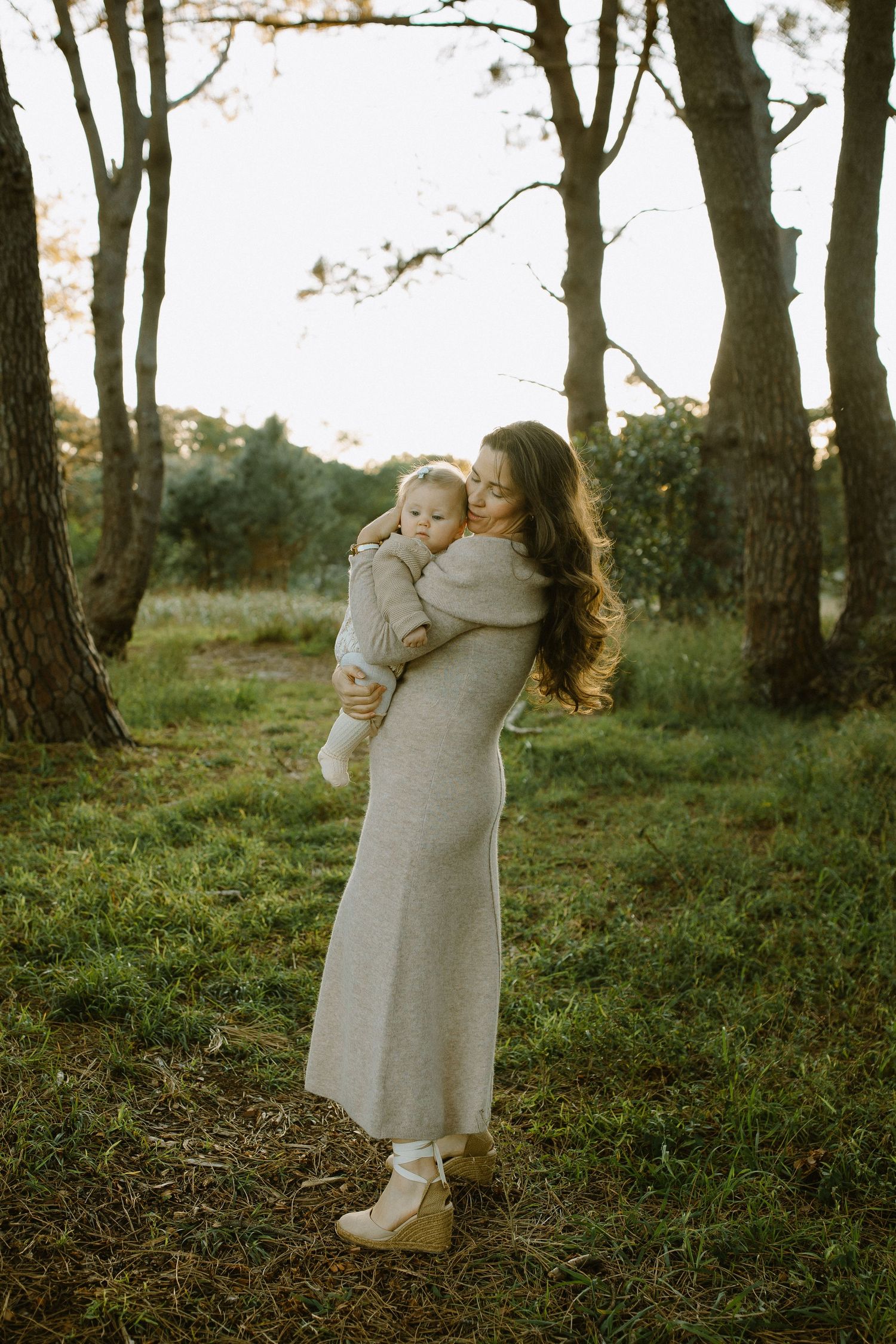 Woman in long grey dress holds baby while standing among trees in natural outdoor setting.