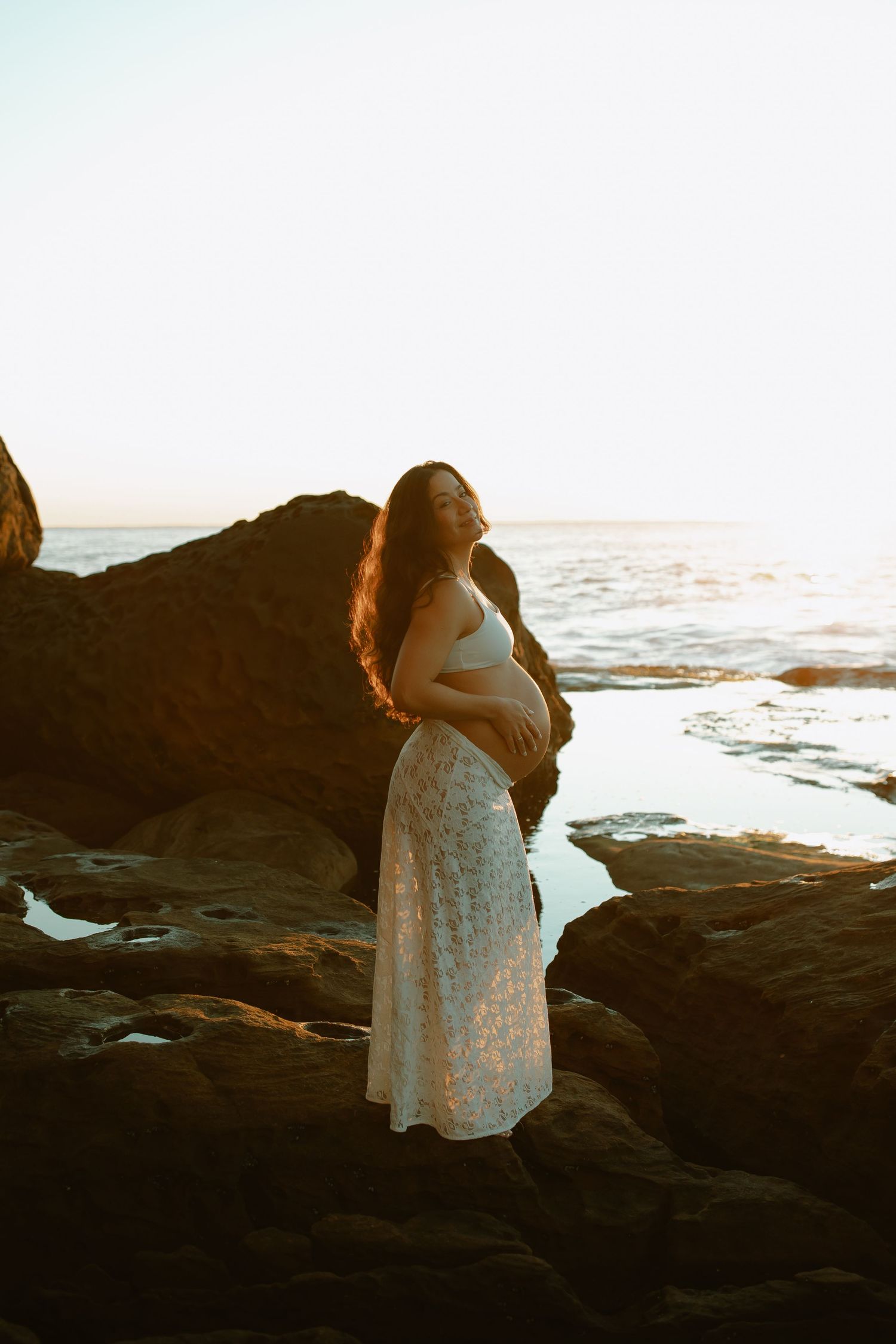 Pregnant woman in white lace dress standing on rocky beach during golden sunset by the ocean.
