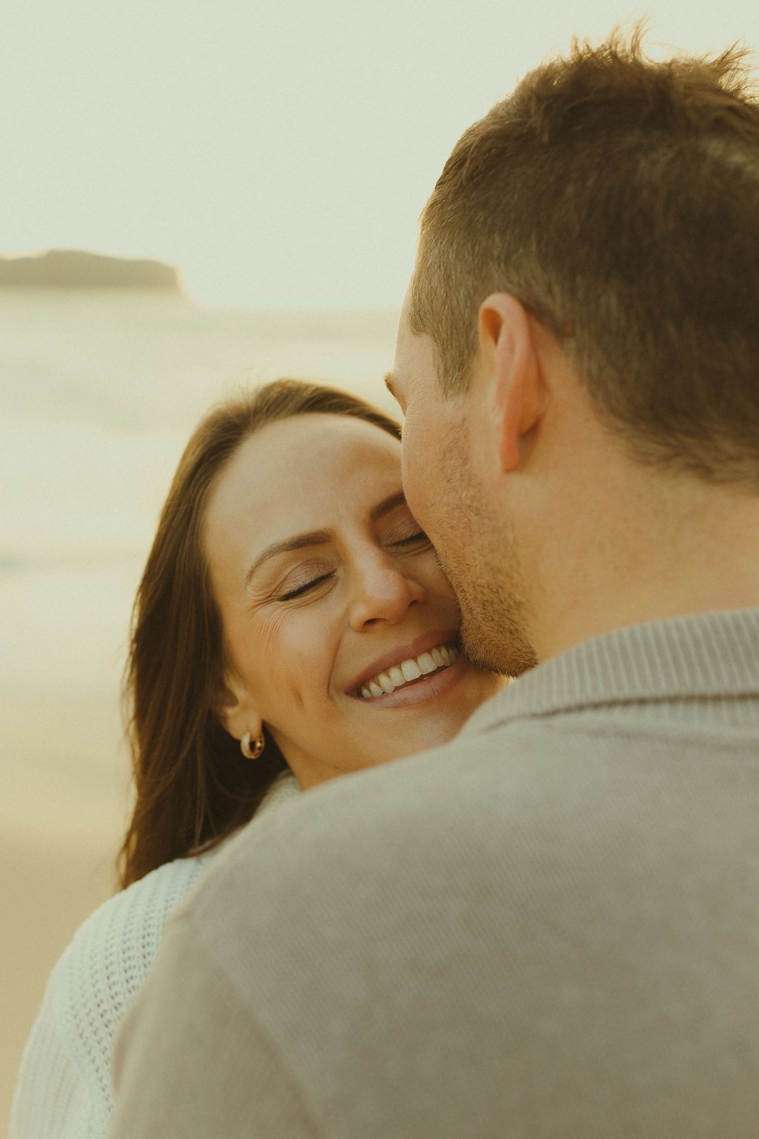 Couple shares an intimate moment during golden hour sunset, with person smiling warmly while embracing partner.